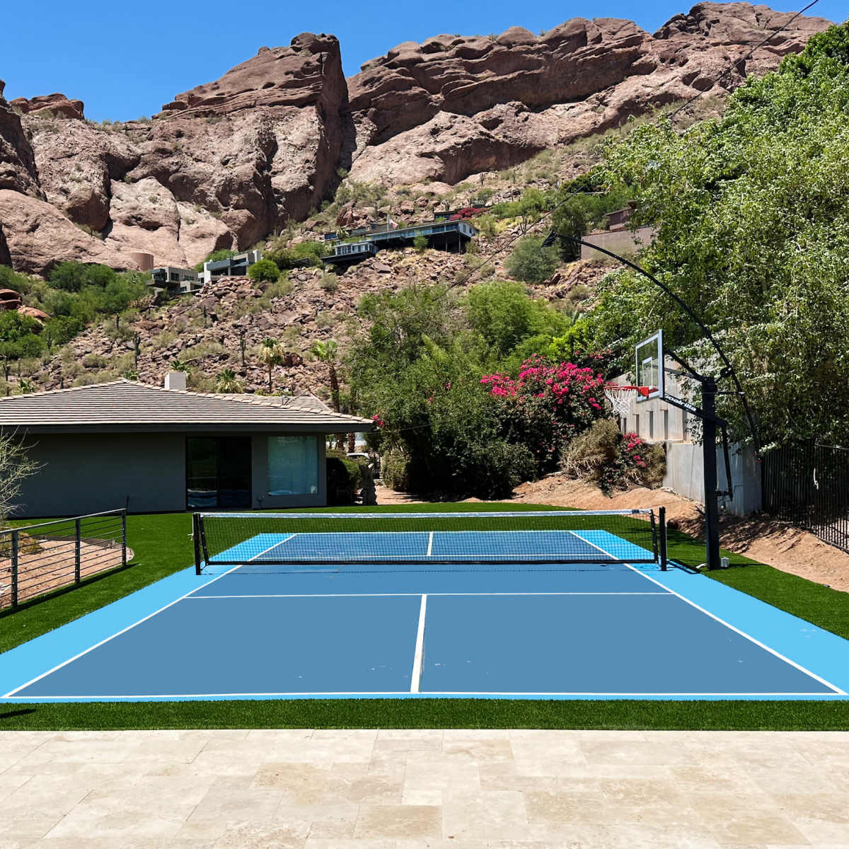 A tennis court with synthetic blue surface surrounded by rocky hills and a house in the background.