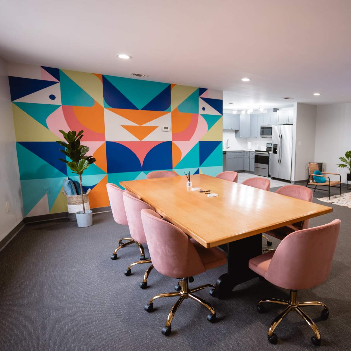 A modern meeting room with a large wooden table surrounded by pink chairs, a colorful geometric mural on one wall, and a comfortable lounge area with sofas and a television.