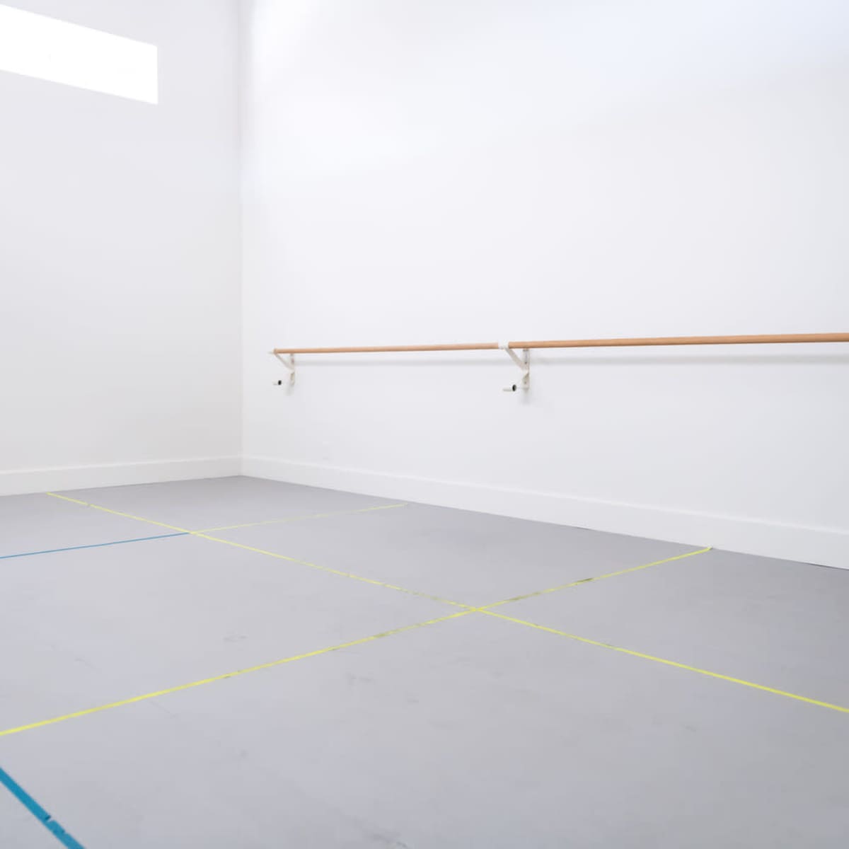 A dance studio with wooden ballet bars mounted on a white wall and a marked floor featuring blue and yellow lines.