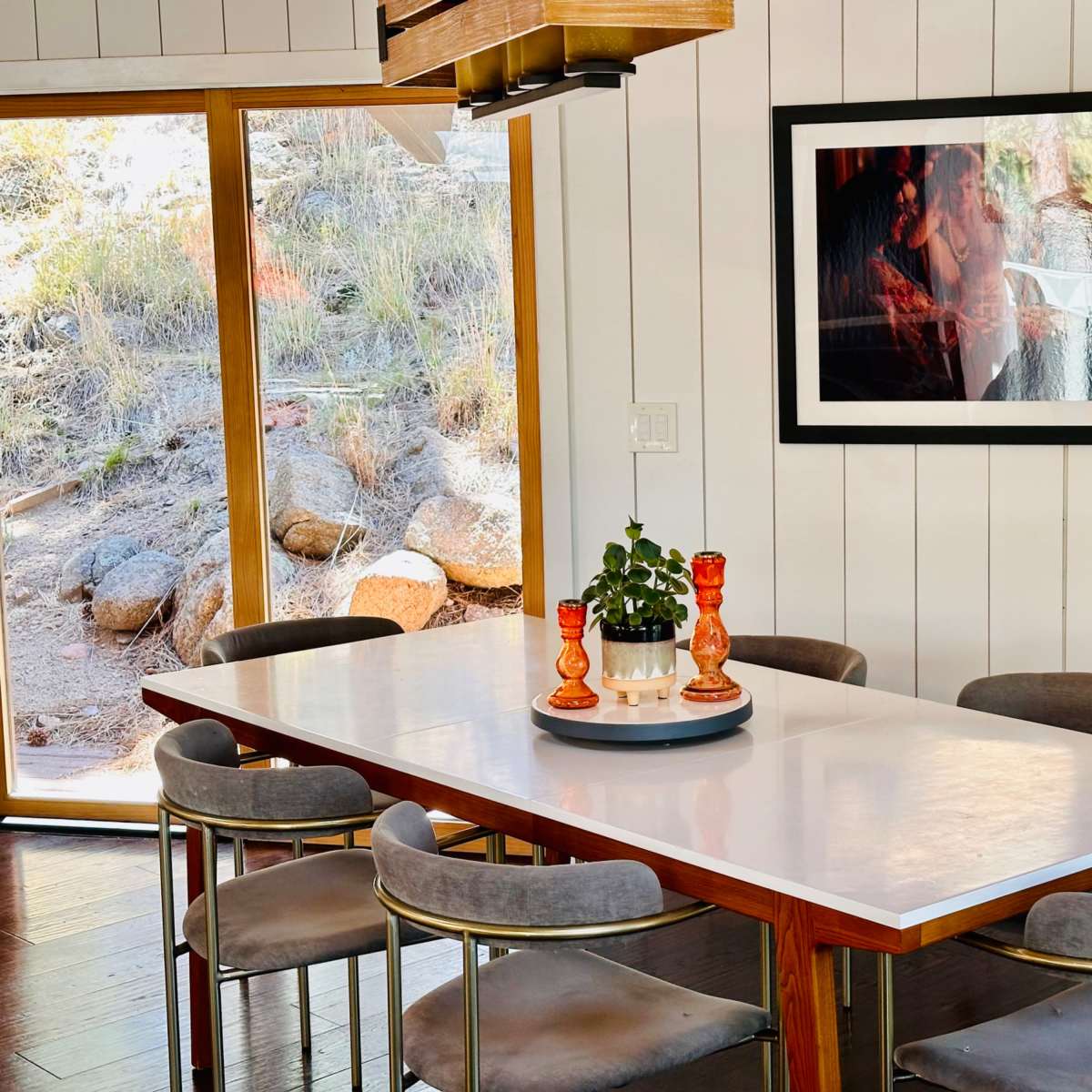 A modern dining area features a white table surrounded by gray chairs, with a large window displaying a rocky outdoor scene and a framed photo on the wall.