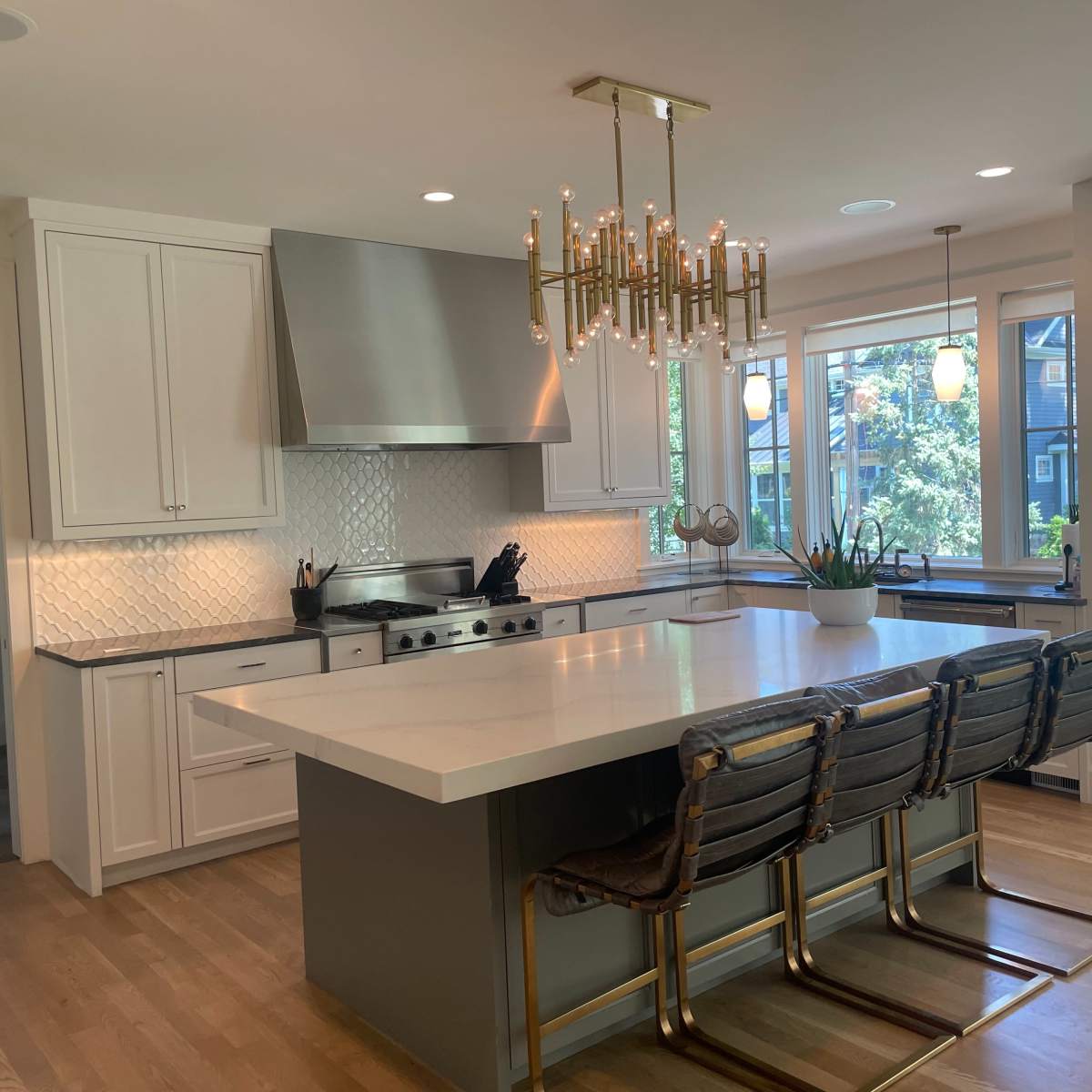 A modern kitchen featuring white cabinetry, a large island with bar seating, a stainless steel range hood, and bright windows overlooking a garden.