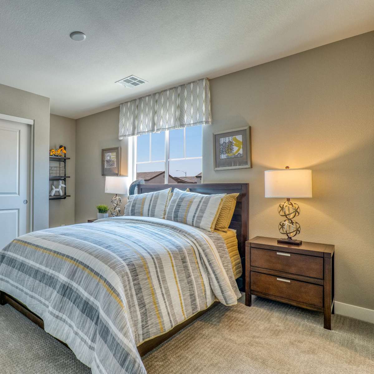 A neatly arranged bedroom featuring a bed with a striped blanket, a nightstand with a lamp, and a window with a patterned valance.