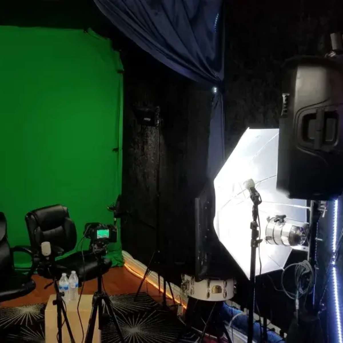A video production setup featuring two black chairs in front of a green screen, surrounded by lighting equipment and a camera on a tripod.