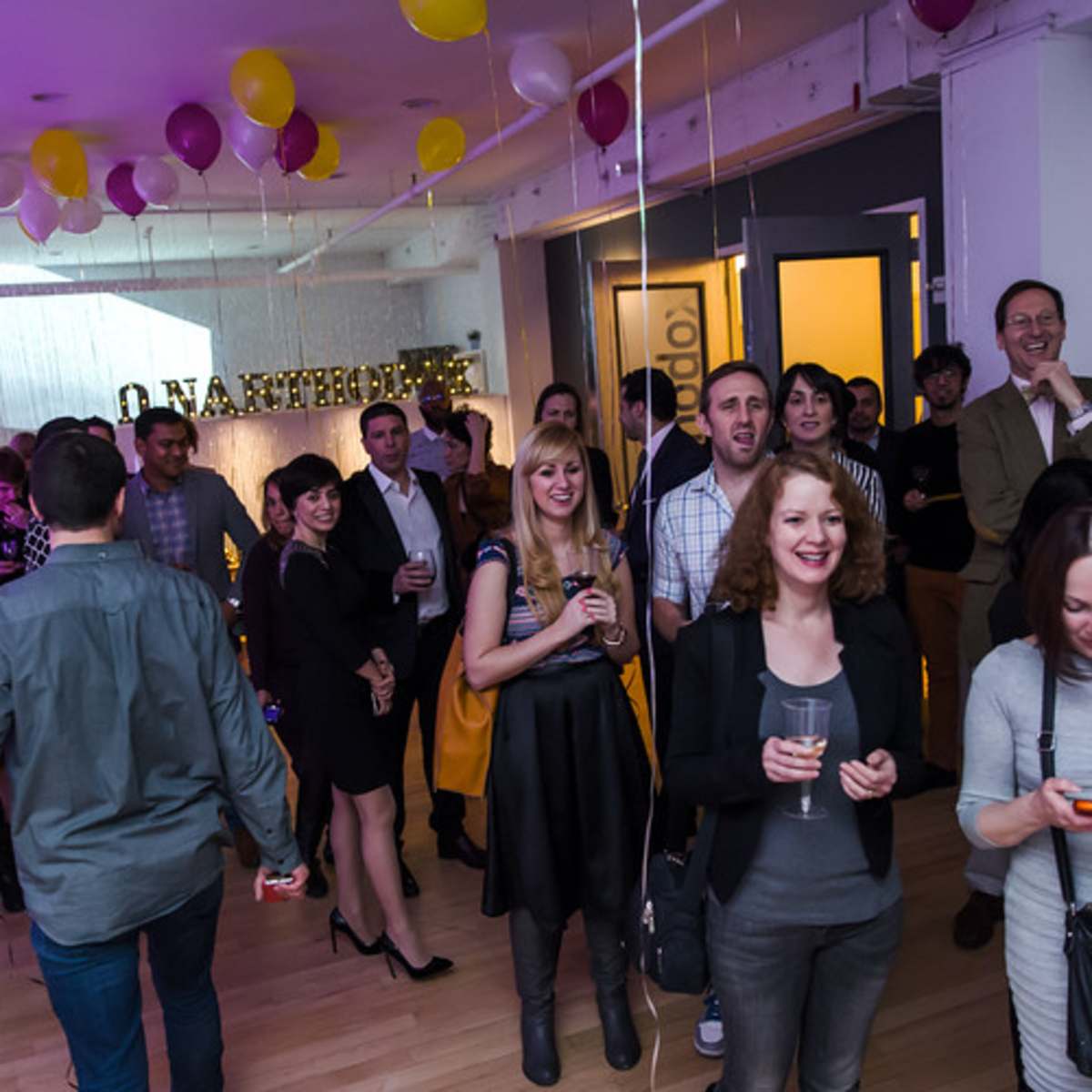 A crowd of people gathers in a brightly lit room decorated with balloons, chatting and holding drinks during an event.