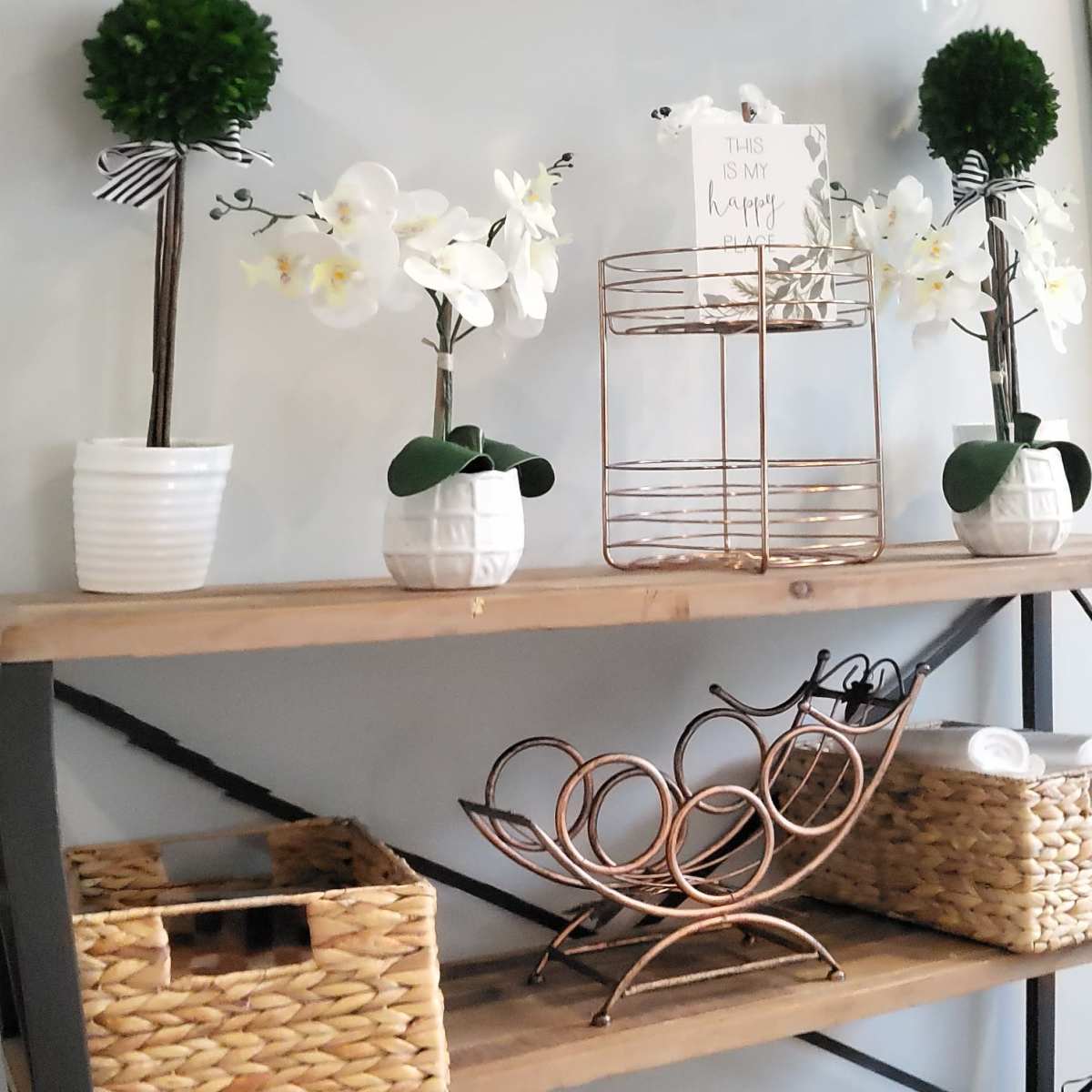 A wooden shelving unit displays various decorative items, including plants, glassware, and storage baskets against a light wall.
