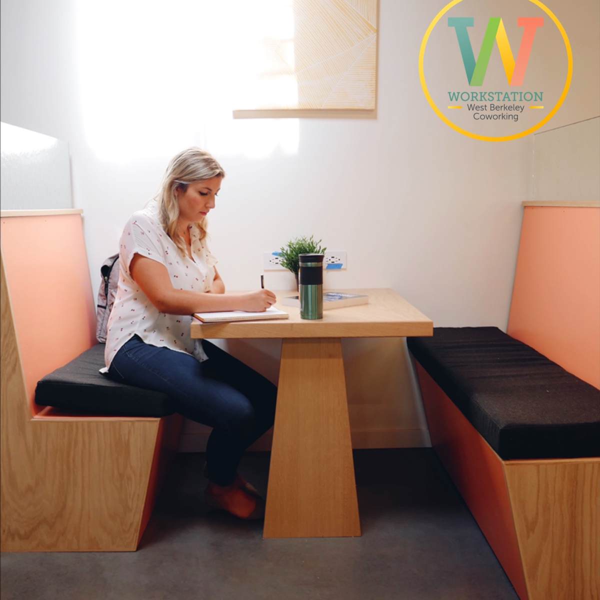 A woman sits at a wooden table in a modern coworking space, writing in a notebook while surrounded by cozy seating.