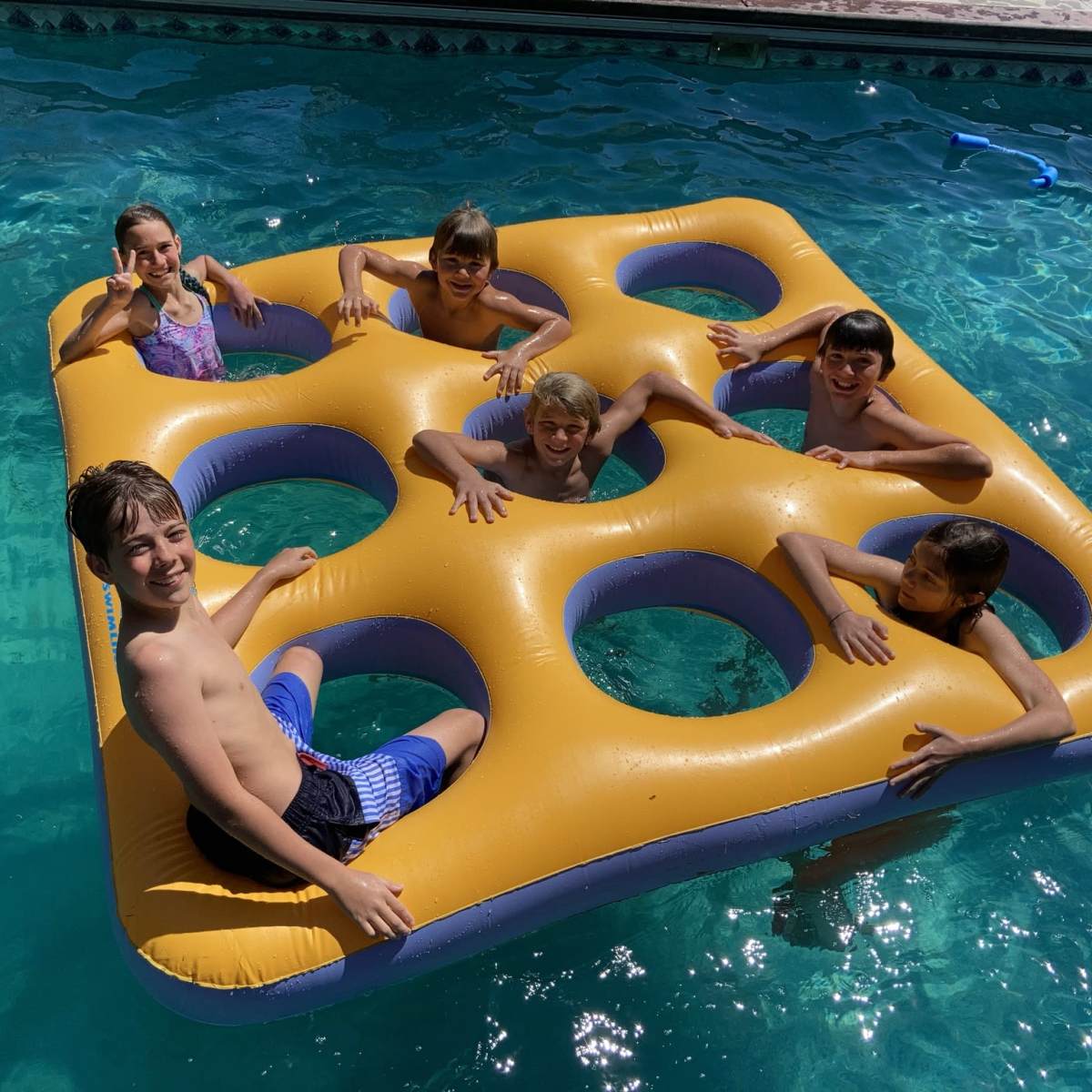 A group of six children is playing on a large, yellow square float with holes in a swimming pool.