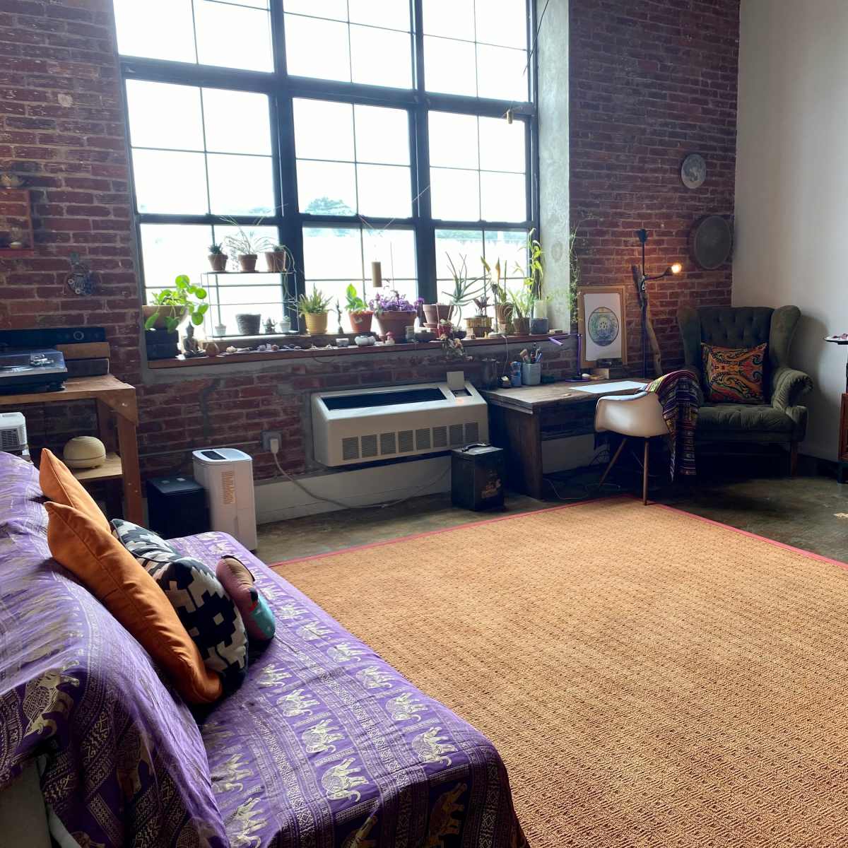 A spacious living room with exposed brick walls, a large window, a couch with colorful cushions, a wooden desk, and various plants placed around the room.