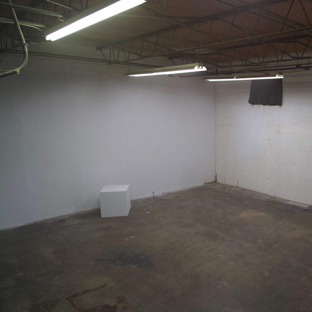 A sparse, empty industrial space with a white wall, a concrete floor, and a single white pedestal positioned against one wall.