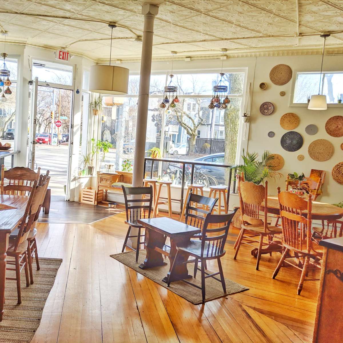A cozy café interior with wooden tables and chairs, decorated with woven wall art and large windows overlooking a street.