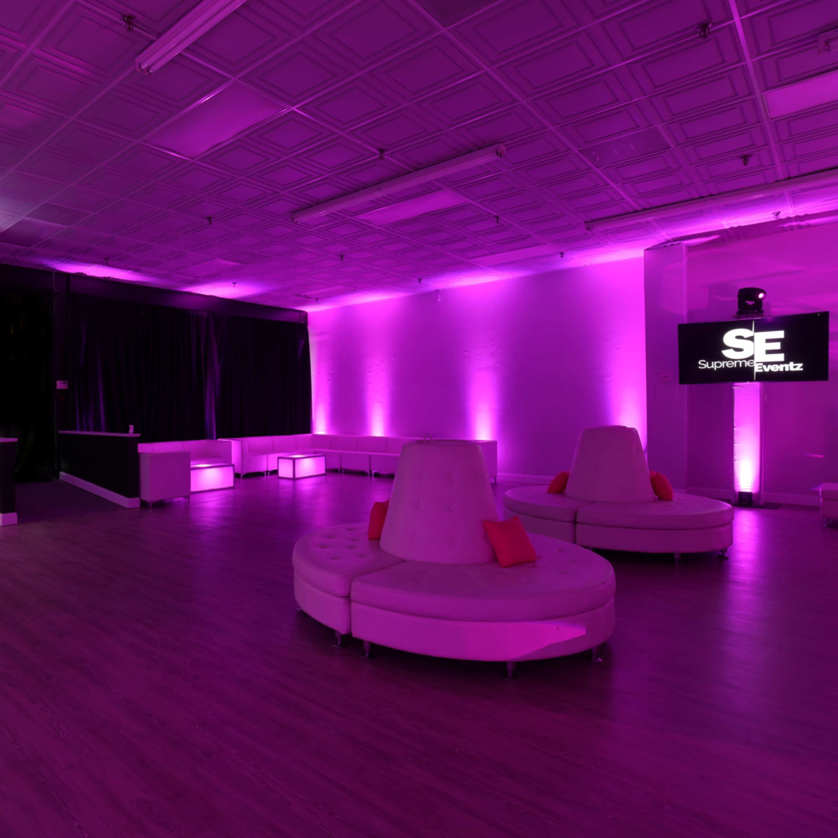 The room features a spacious layout with purple lighting, circular white seating, and a simple bar setup against a black backdrop.