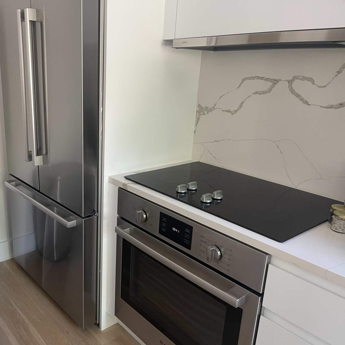 A modern kitchen corner featuring a stainless steel refrigerator, a built-in oven and cooktop, and a garbage can.