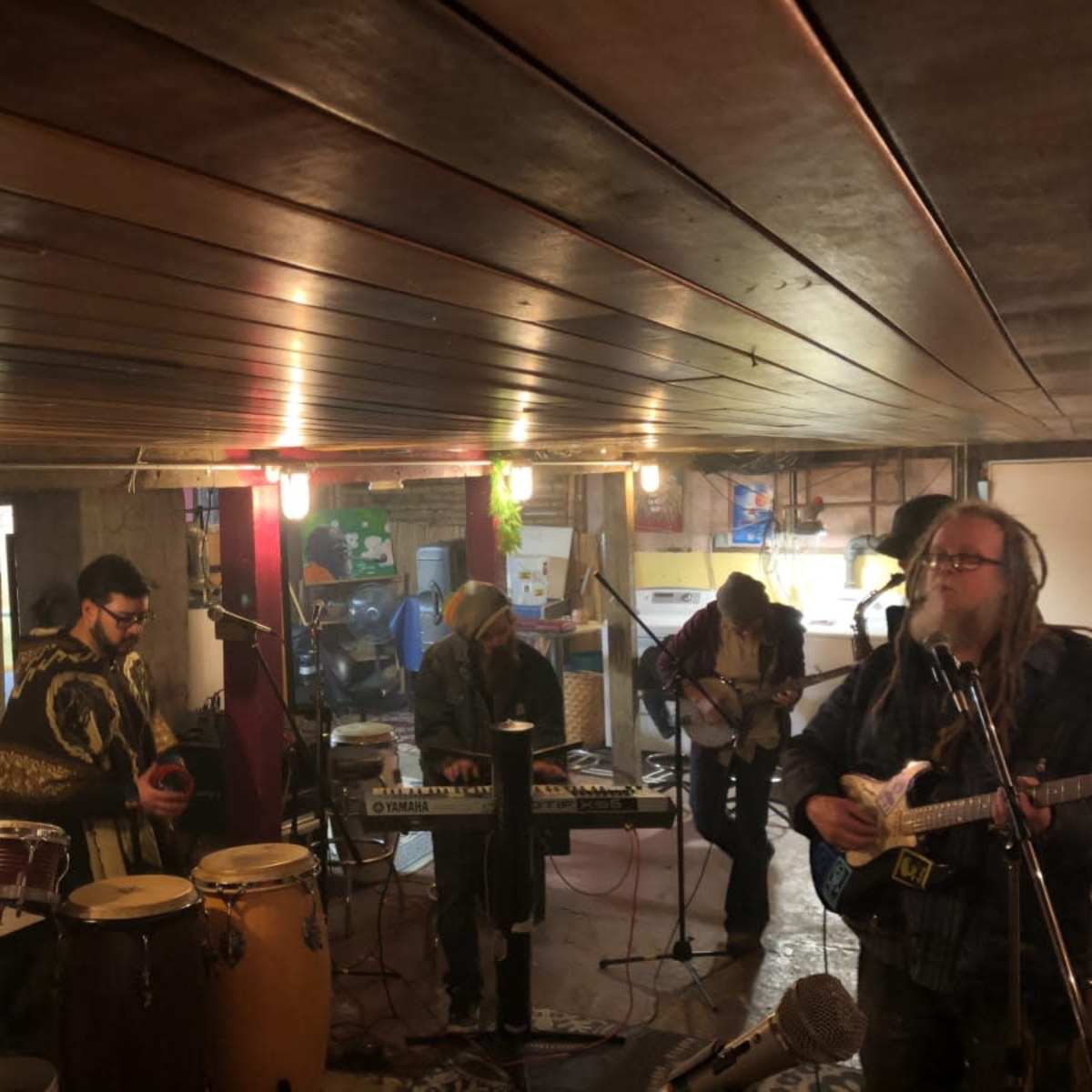 A group of musicians plays various instruments in a dimly lit basement studio.