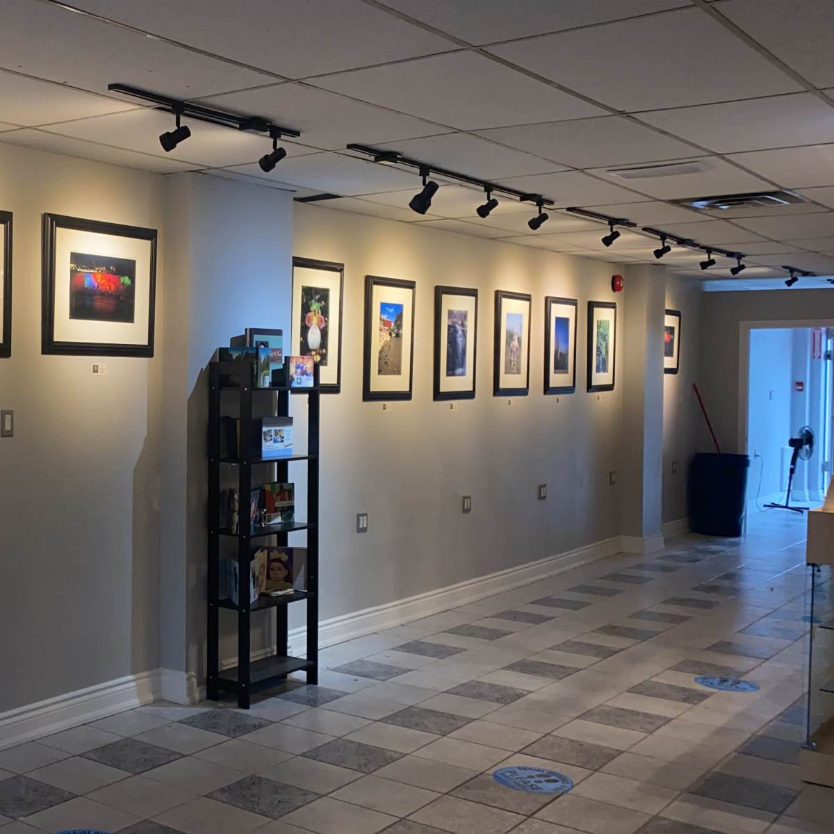 A gallery corridor featuring a row of framed artworks displayed on the wall, accompanied by a black shelving unit on the right.
