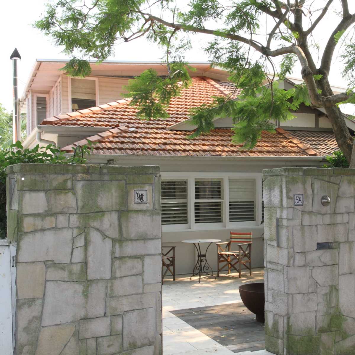 A single-story house with a tiled roof is visible through a stone and wooden gate, accompanied by outdoor seating beneath a tree.