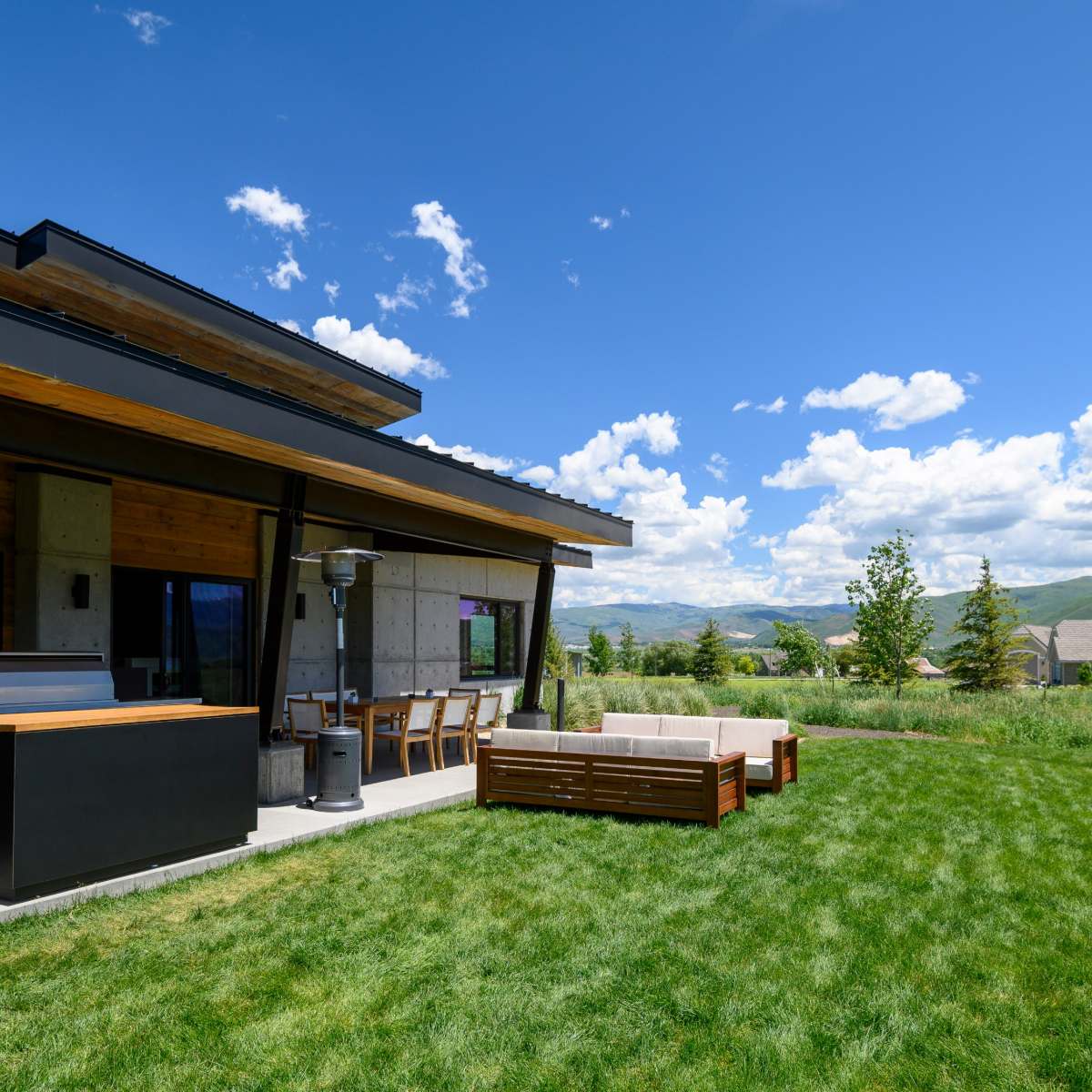 A modern house with a patio area featuring outdoor seating and a grill overlooks a green grassy landscape under a blue sky with clouds.