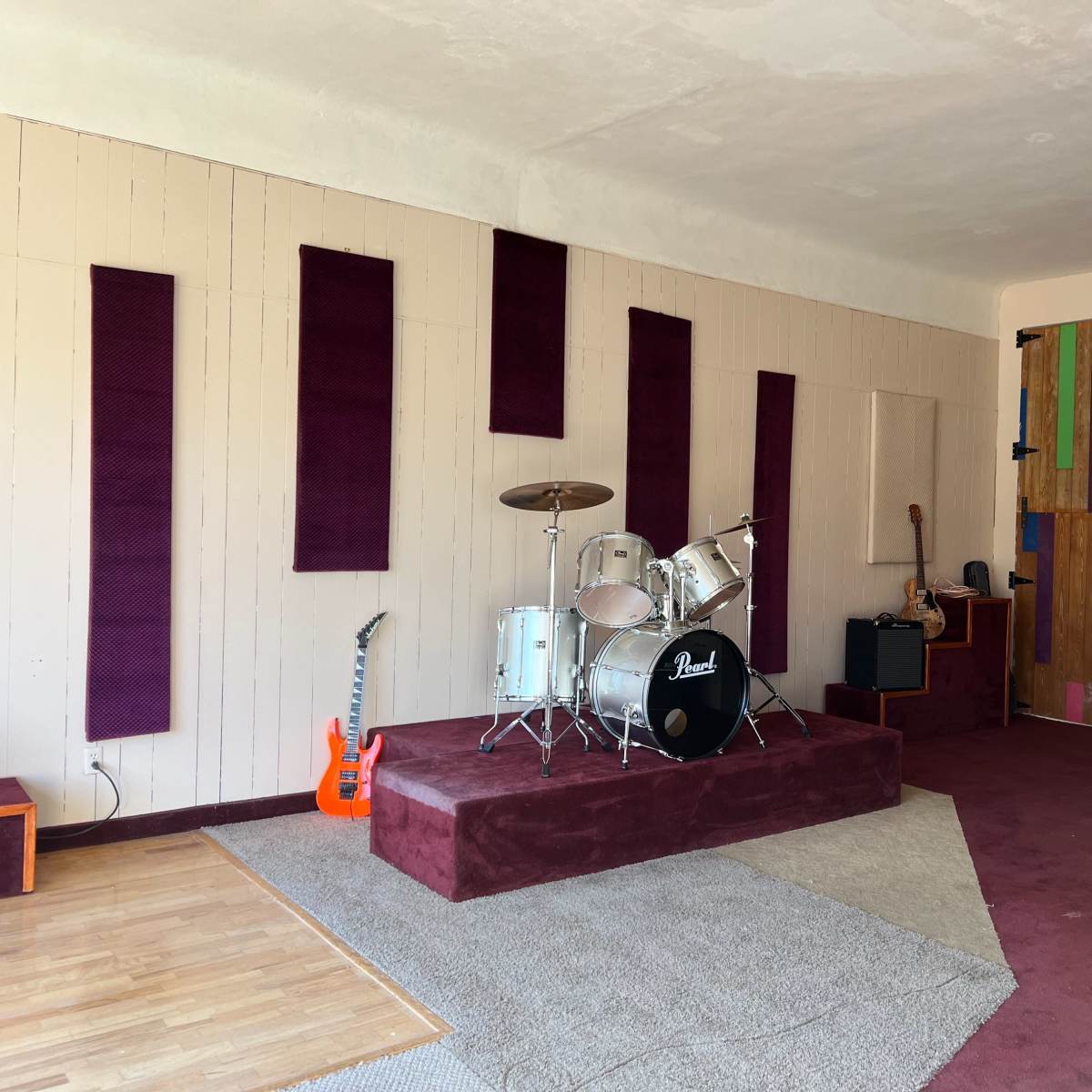A spacious room designed for music practice, featuring a drum set on a raised platform, an electric guitar, and a colorful wooden door.