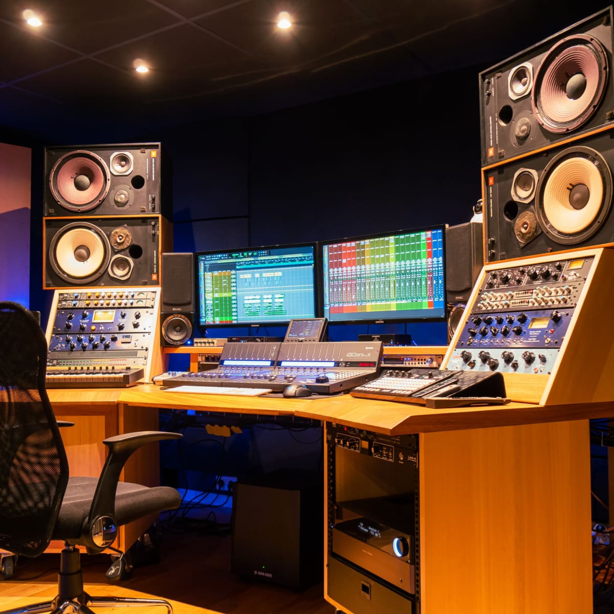 A modern recording studio with a mixing console, monitors, and audio equipment arranged around a central workstation.