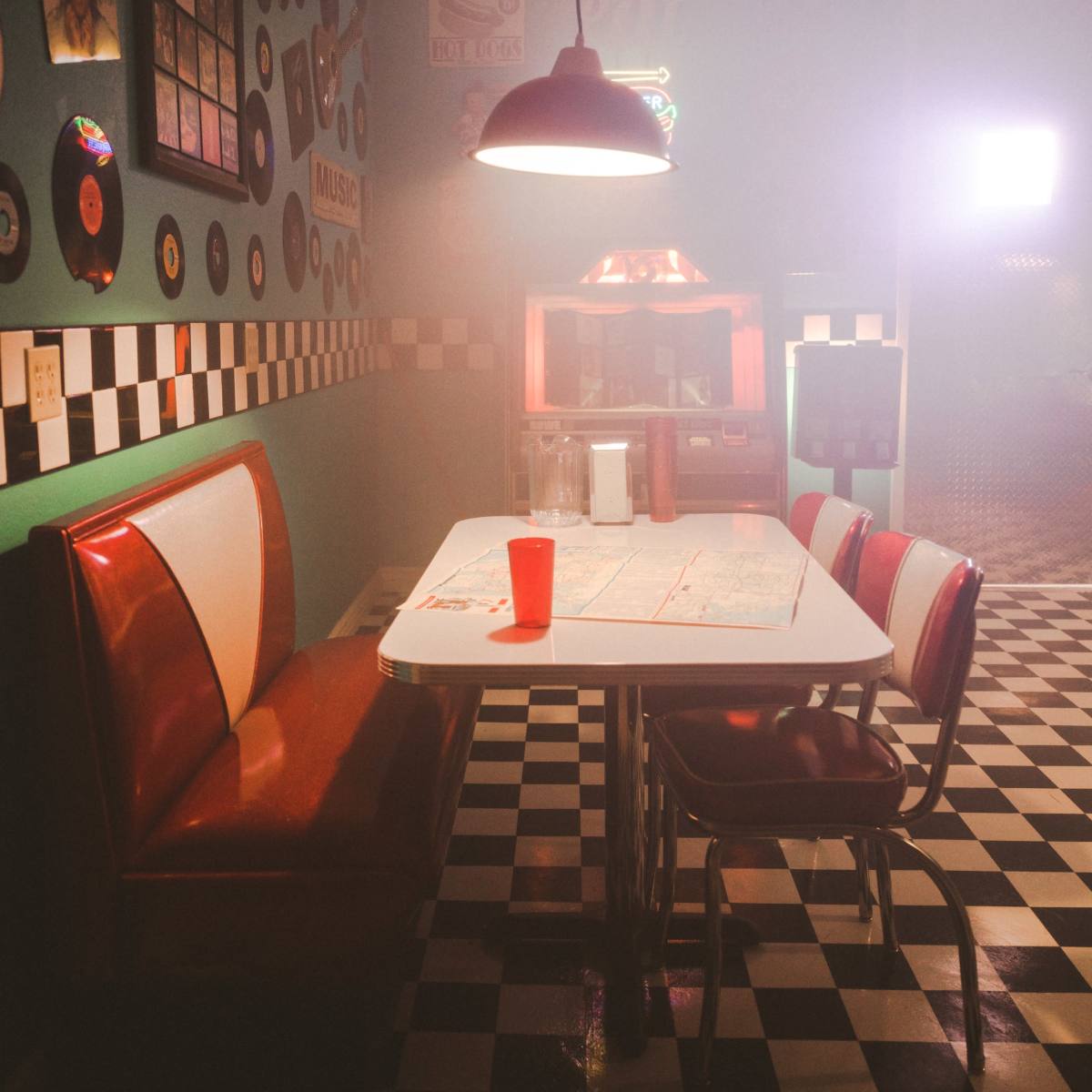 A vintage diner scene features a red booth and a white table set on black-and-white checkered flooring, illuminated by a hanging light, with a red cup on the table.