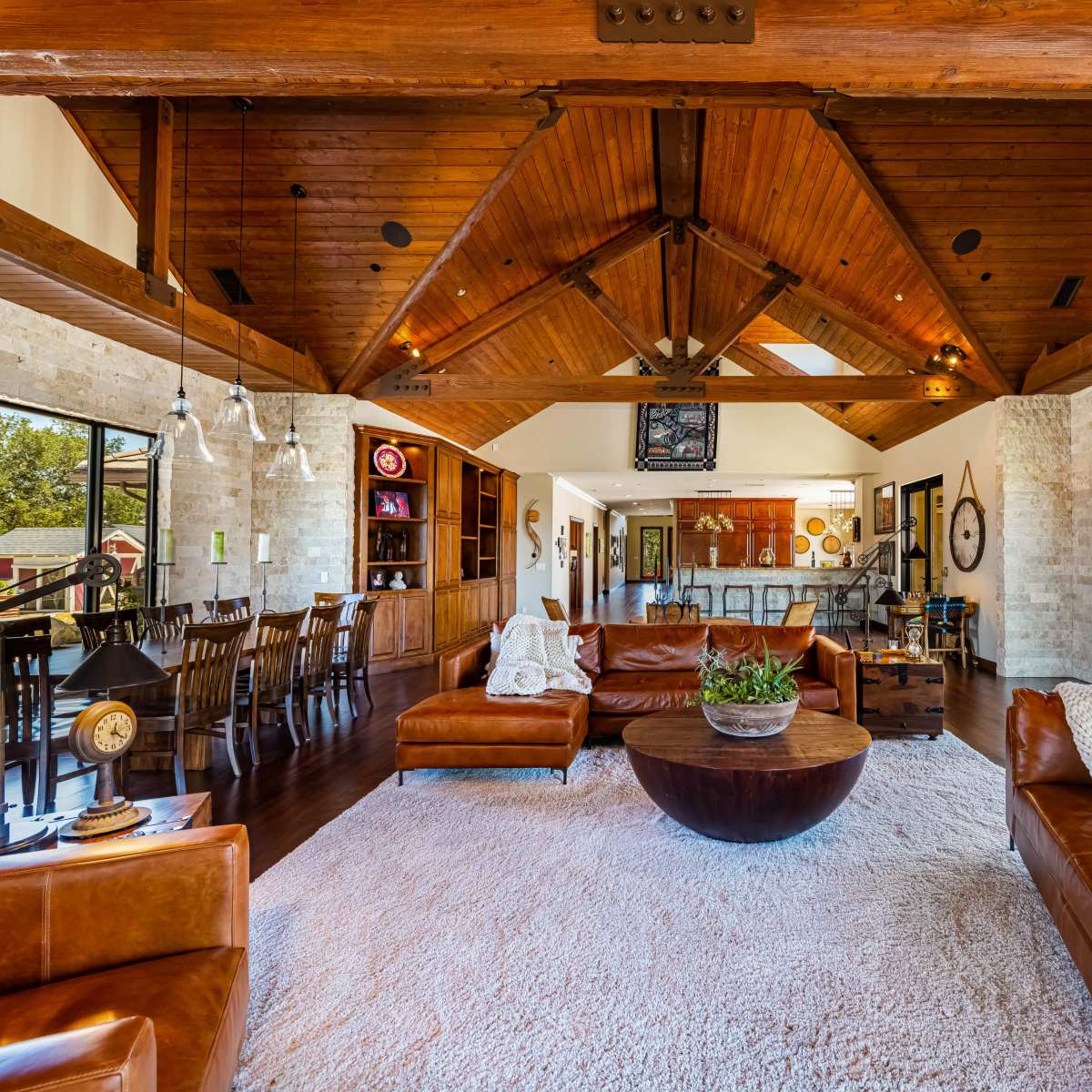 A spacious living room with exposed wooden beams, large windows, leather sofas, and a round coffee table on a light rug.