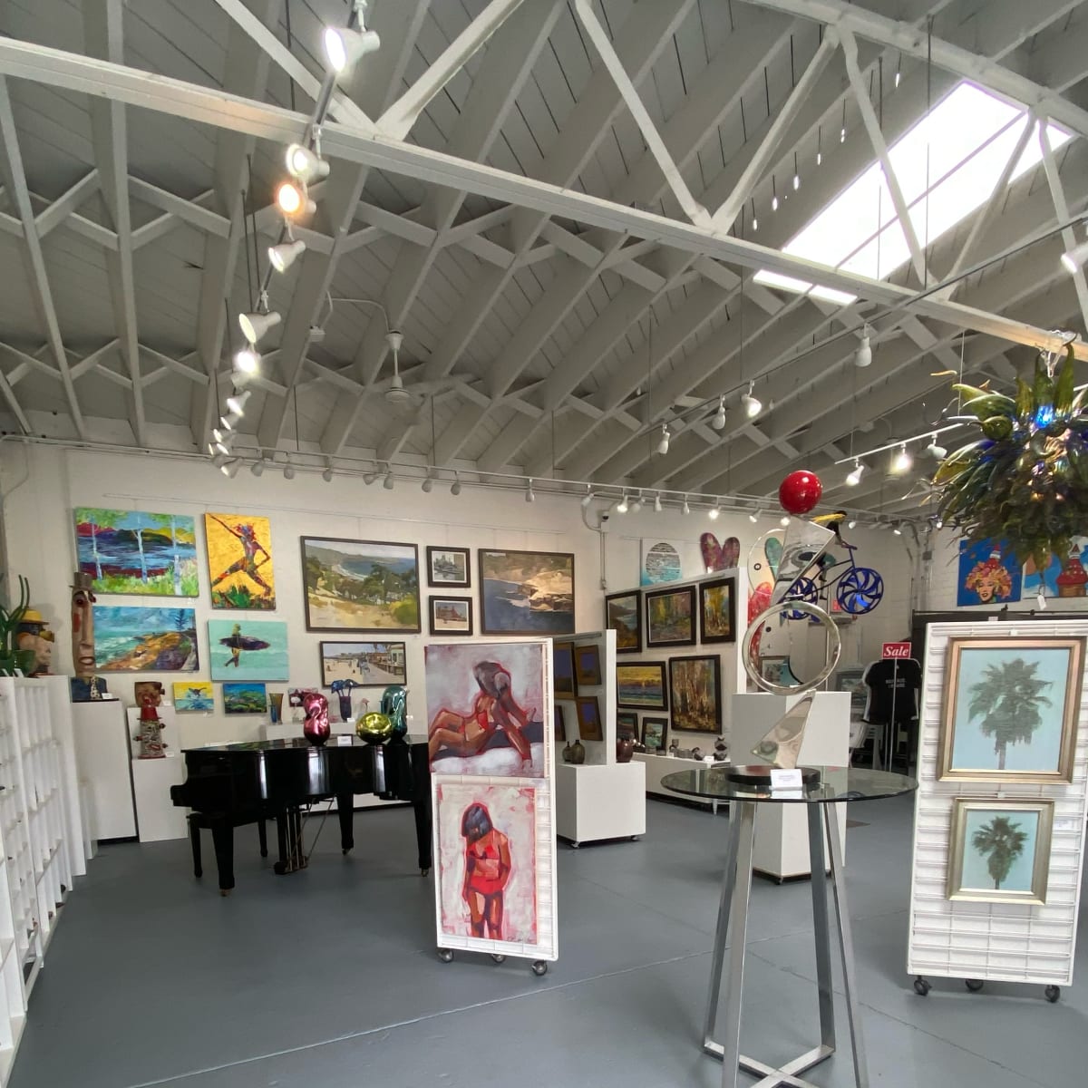 An indoor art gallery featuring various paintings and sculptures displayed on white stands, with a black piano in the corner.