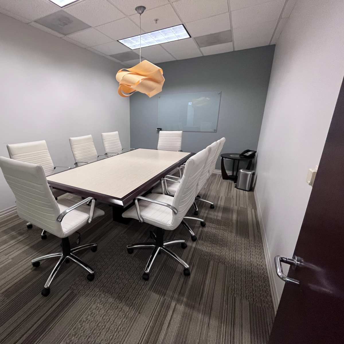 A modern conference room features a long rectangular table surrounded by eight white swivel chairs, with a pendant light hanging above.