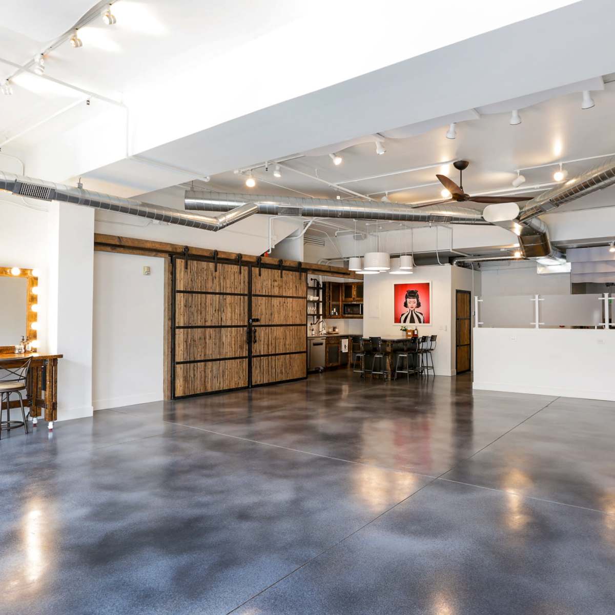 A spacious, modern room featuring a polished concrete floor, exposed metal ductwork, and a kitchenette with a sliding barn door.