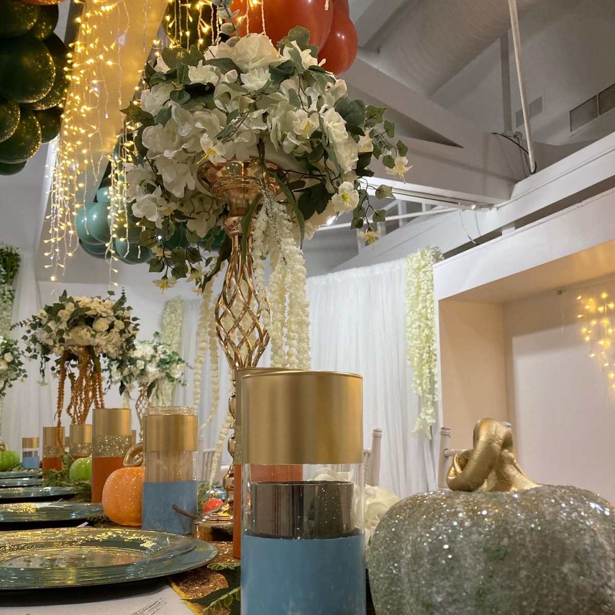 A elegantly arranged dining table with decorative centerpieces, including vases with flowers and sparkly pumpkins, under a backdrop of fairy lights.