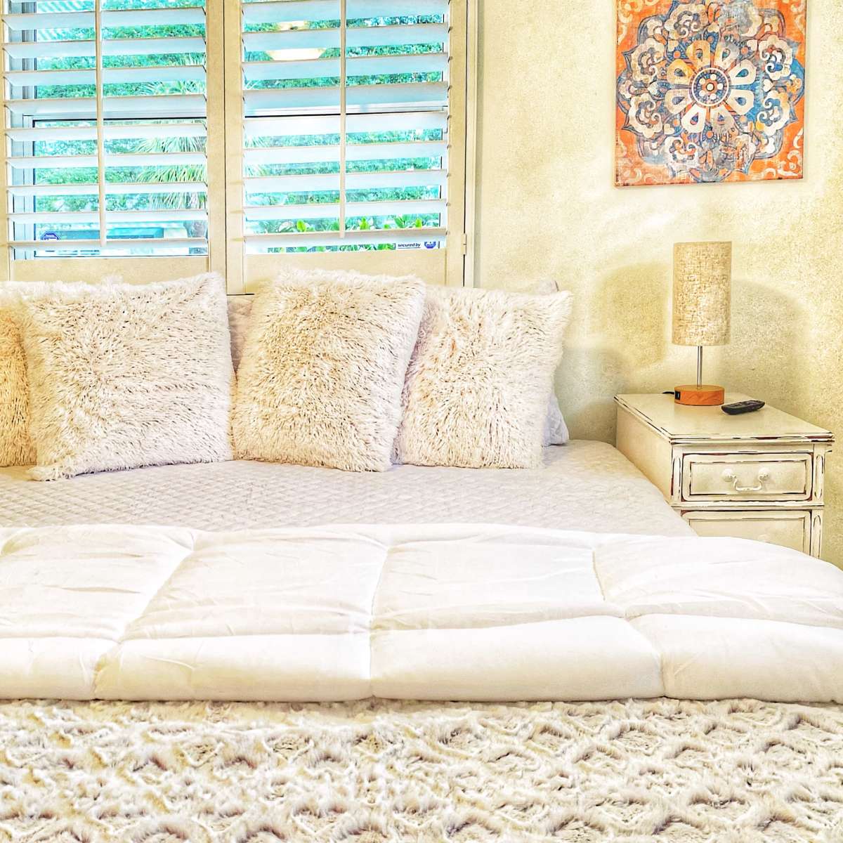A neatly arranged bed with fluffy pillows and a white comforter is positioned against a wall with window shutters and decorative wall art.