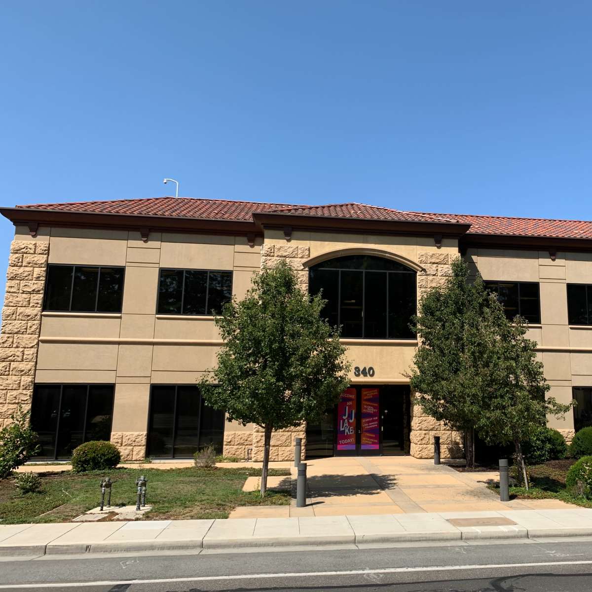 A two-story office building with tan stone exterior and large windows is situated on a landscaped lot.