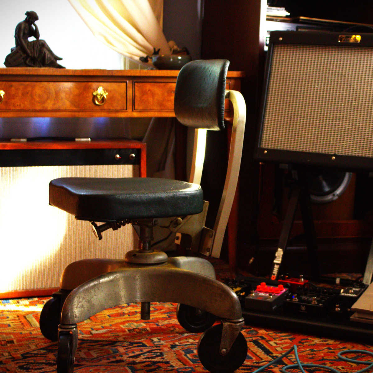 A black office chair is positioned next to a guitar amplifier and effects pedals, surrounded by wooden furniture and textured rugs.