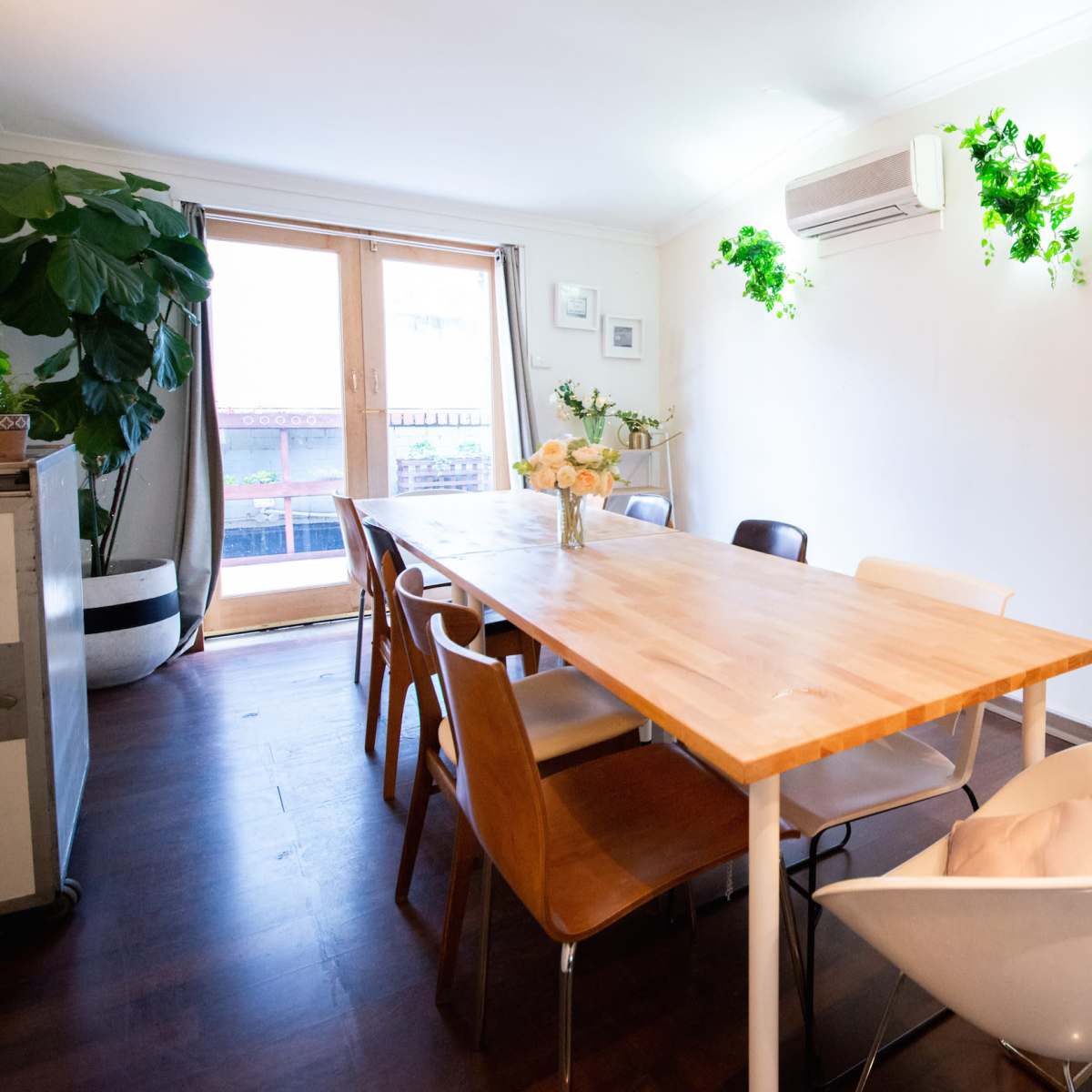 A large wooden dining table is surrounded by various chairs in a well-lit room with potted plants and an air conditioning unit mounted on the wall.