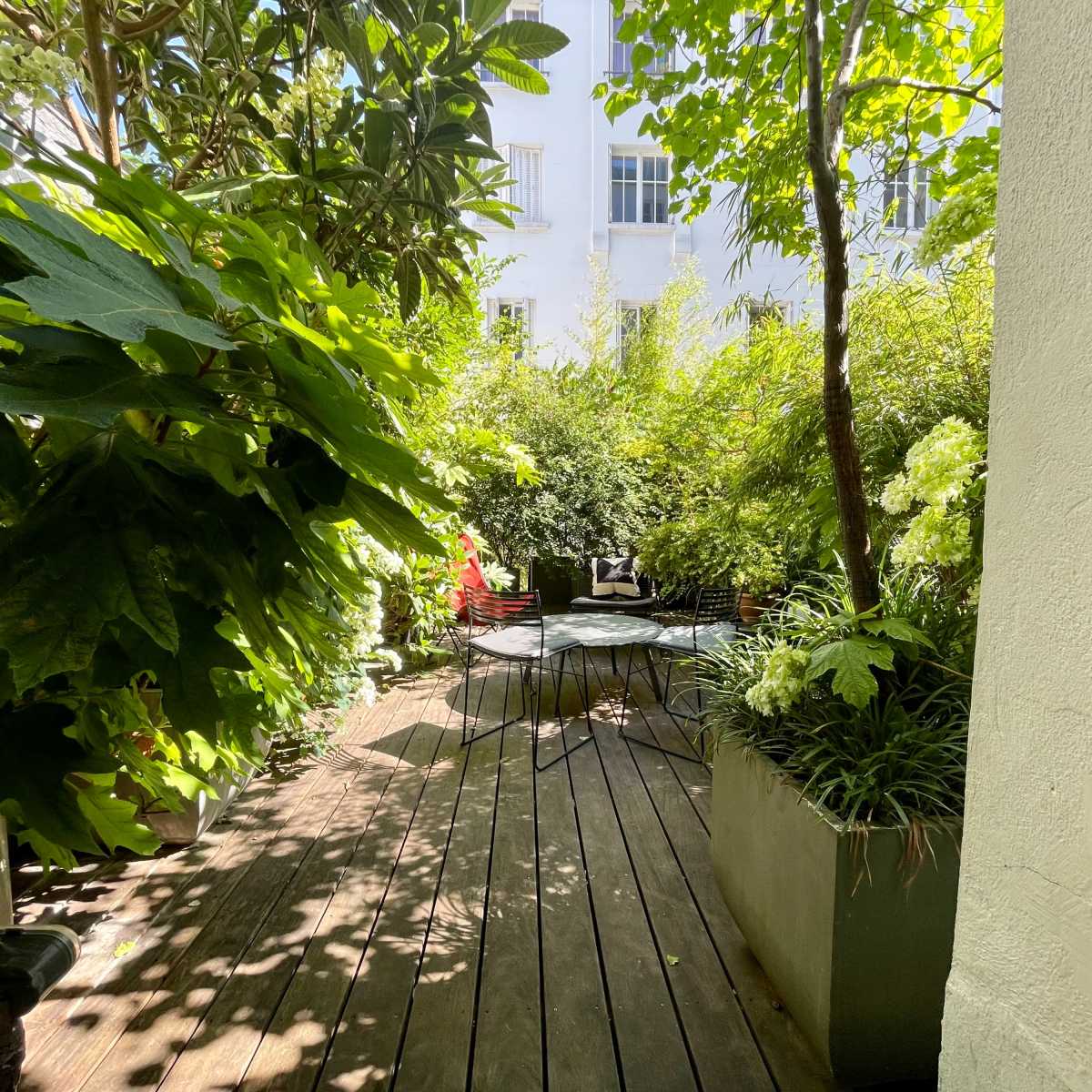 A wooden deck surrounded by lush greenery leads to a small seating area in a garden.