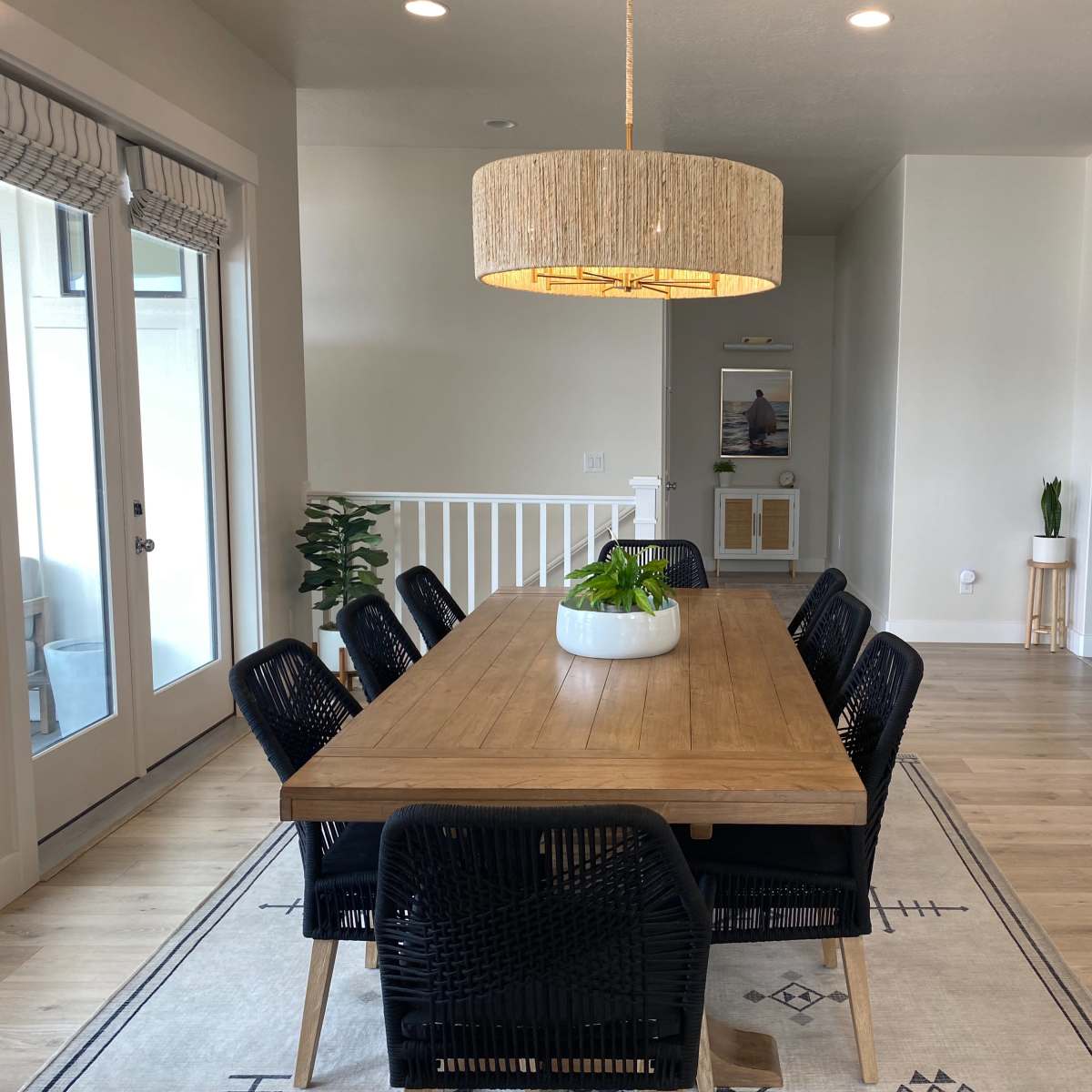 A modern dining area features a wooden table surrounded by six black chairs, with a large pendant light hanging above and a potted plant on the table.