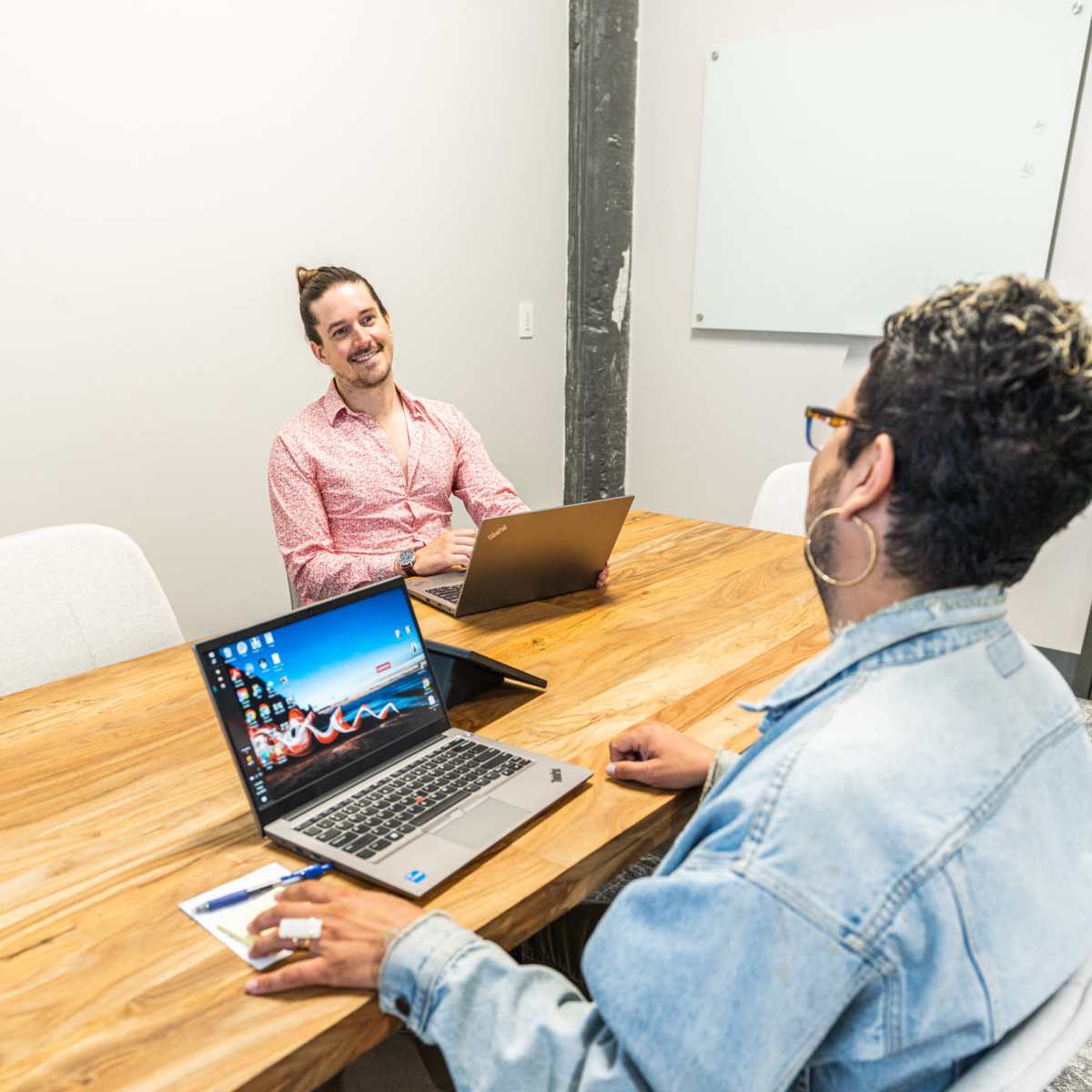 Two people are sitting at a wooden table in a meeting room, each using a laptop and engaging in conversation.