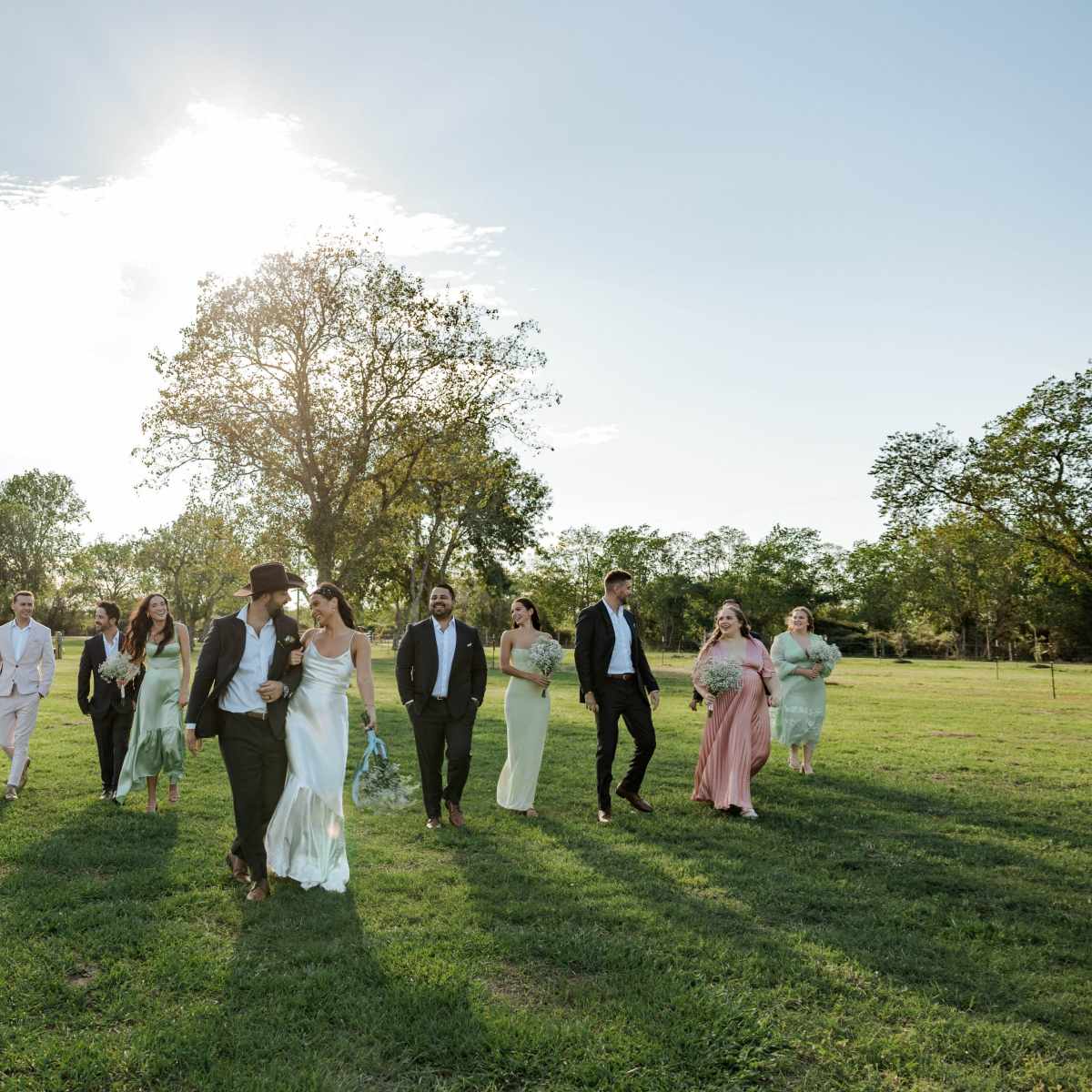 A group of people dressed in formal attire walks across a grassy field with a pond visible in the background.
