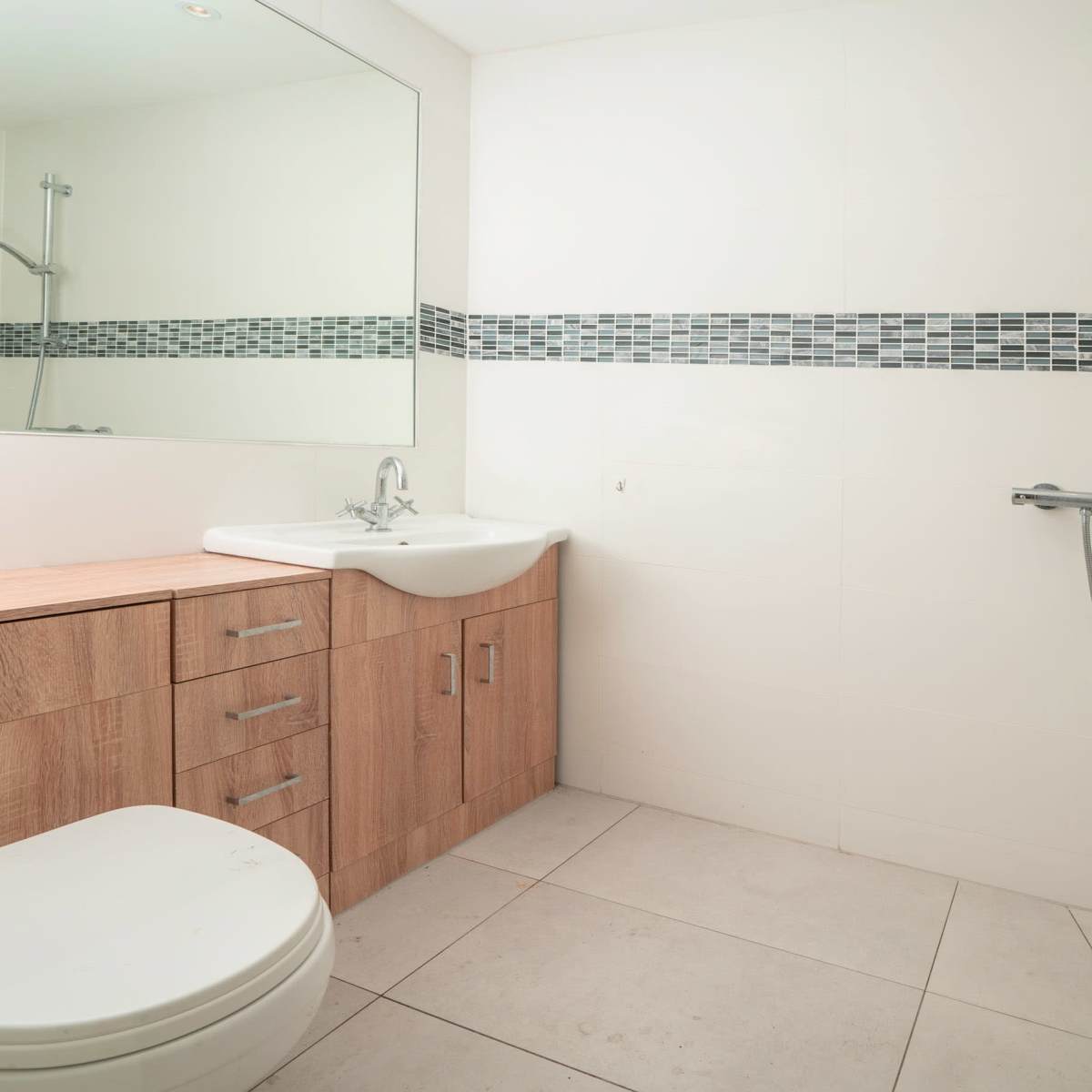 A clean bathroom featuring a shower, sink, wooden cabinet, and tiled floor with a decorative border.