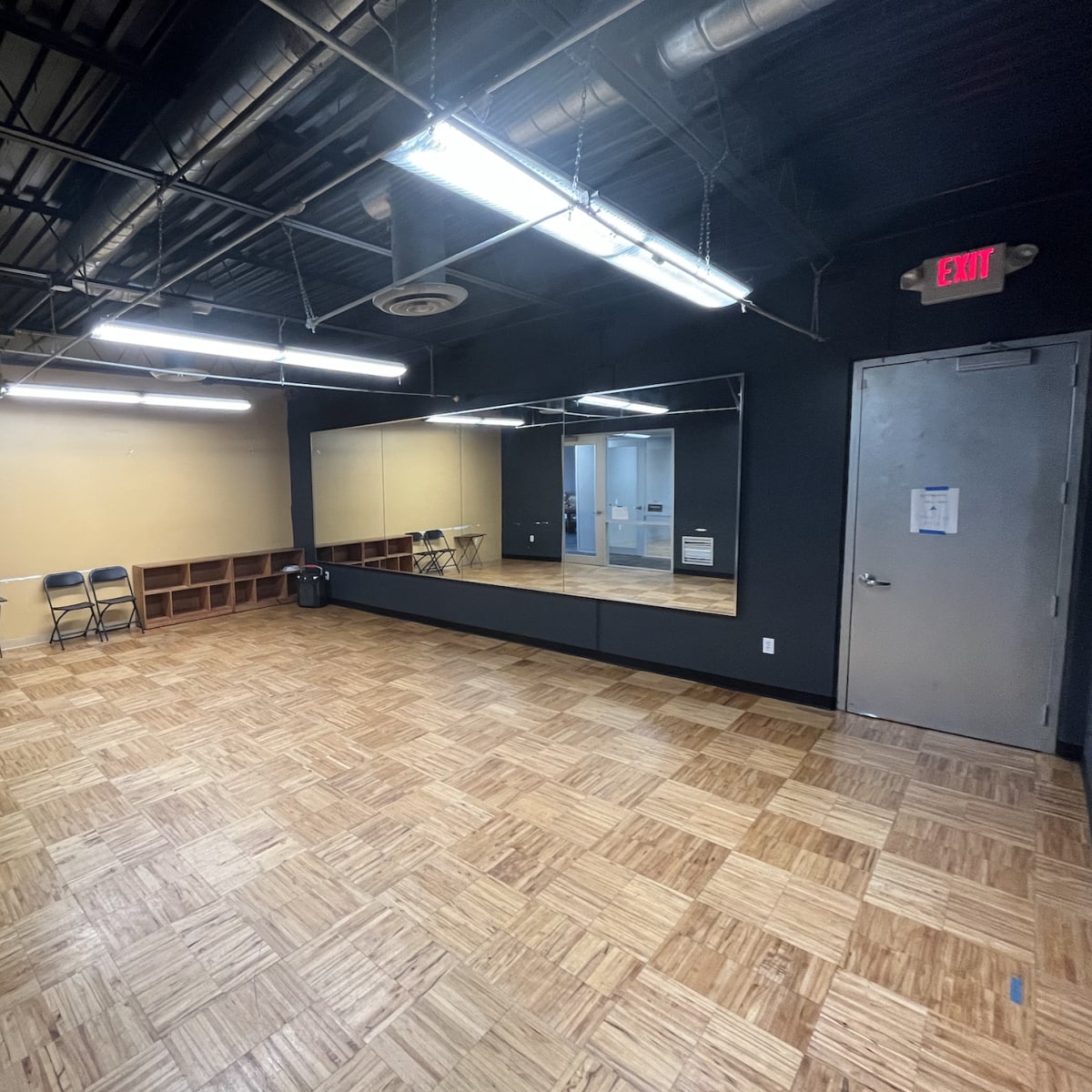 An empty room with a wooden floor, a mirrored wall, and two folding chairs against one wall.