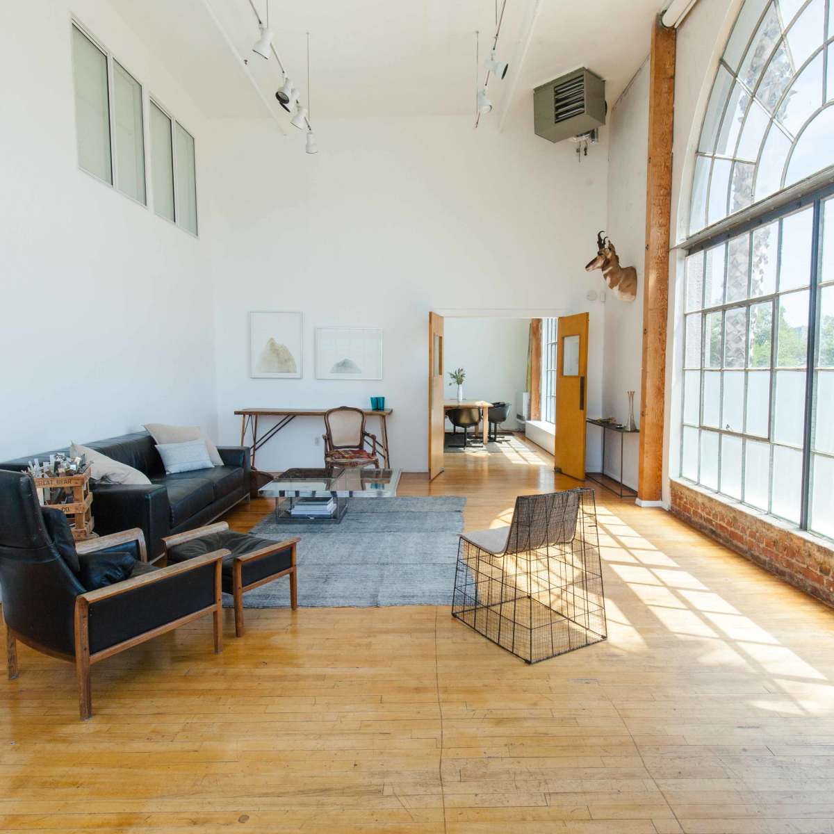 A spacious, brightly lit room with large windows, featuring a seating area with a sofa and two chairs, a painting on the wall, and a dining area in the background.