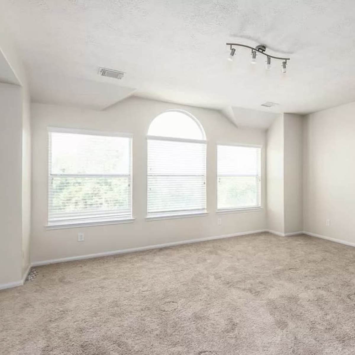 A sparsely furnished room with beige carpet and large windows providing natural light.