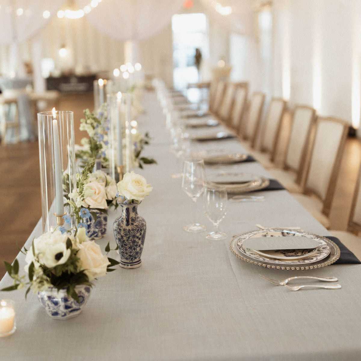 A long, elegantly set dining table is adorned with blue and white vases, candles, and floral arrangements under string lights in a bright, open space.