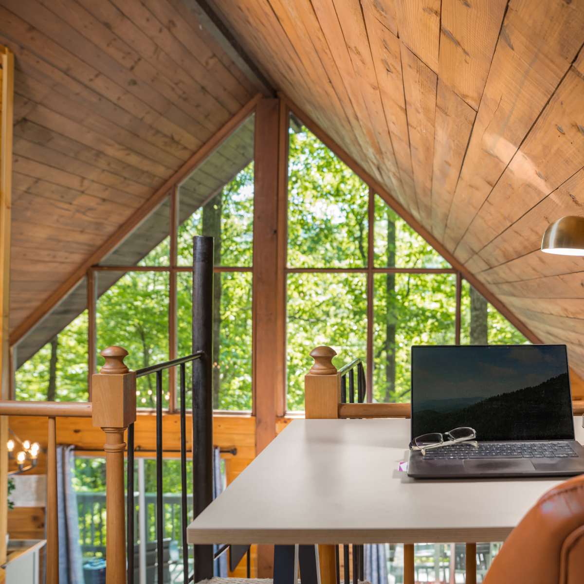A modern workspace is set up in a loft with wooden walls and large windows letting in natural light, featuring a laptop, a desk lamp, and a small plant.
