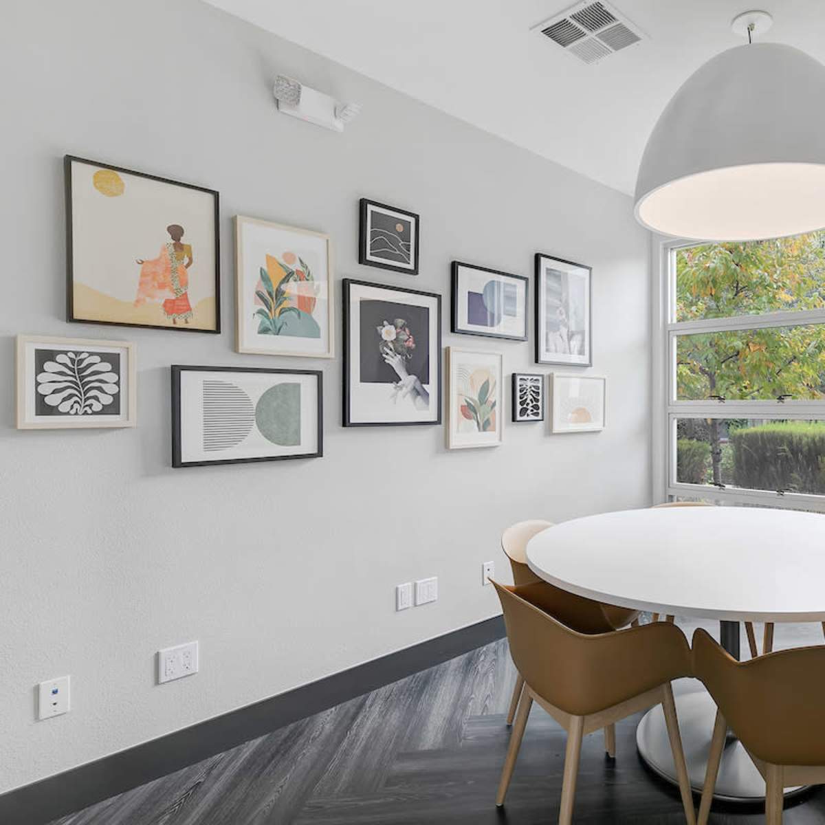 A round white table with four brown chairs is surrounded by framed artwork on a gray wall, with a large window and a plant in the corner.