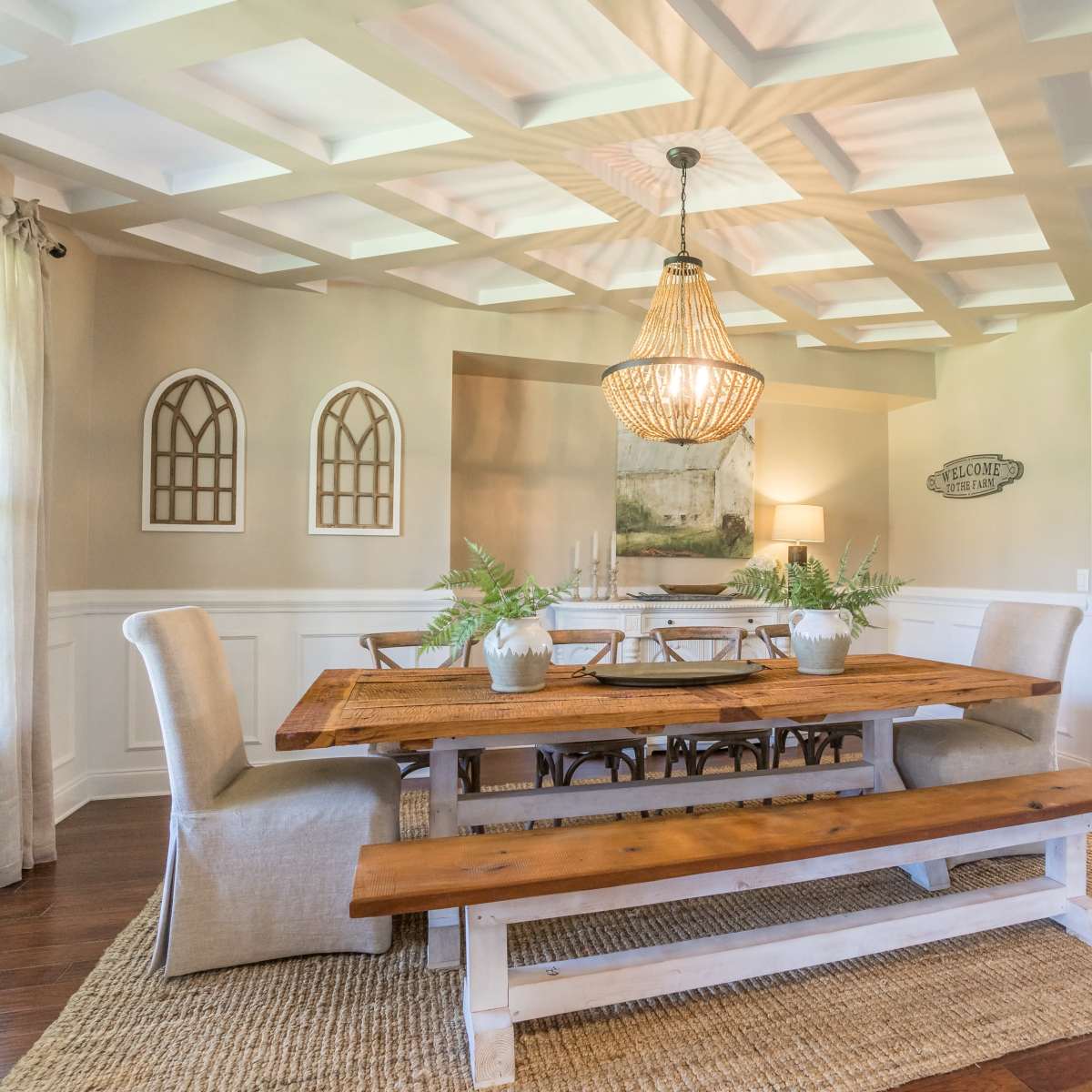 A spacious dining room features a wooden table with benches, a chandelier above, and decorative wall accents, with a doorway leading to a kitchen.