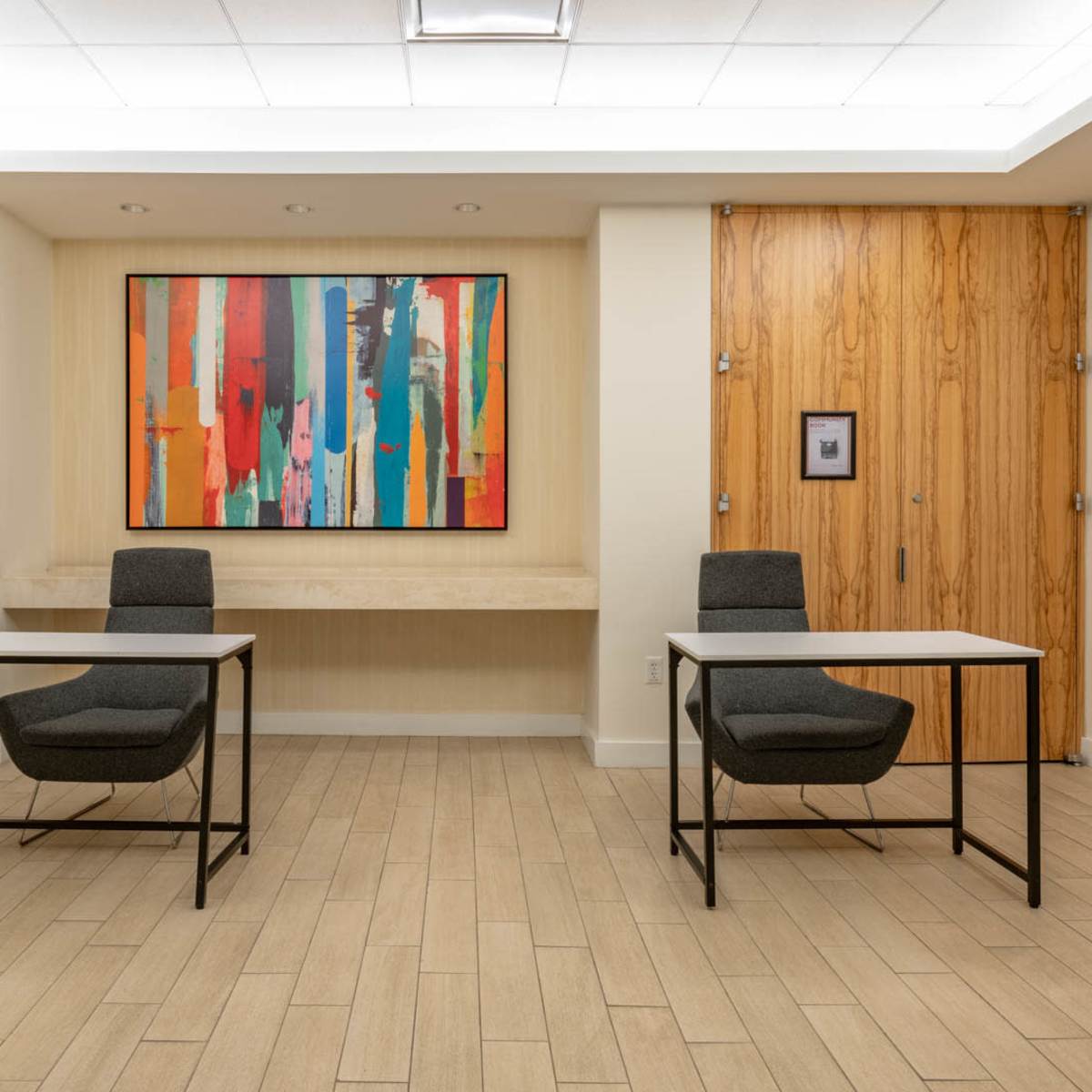 A minimalist room with two chairs and tables facing a colorful abstract painting on the wall.