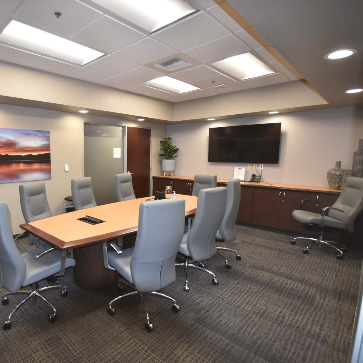 A modern conference room equipped with a large wooden table surrounded by ten gray swivel chairs, along with a wall-mounted television and a landscape painting.