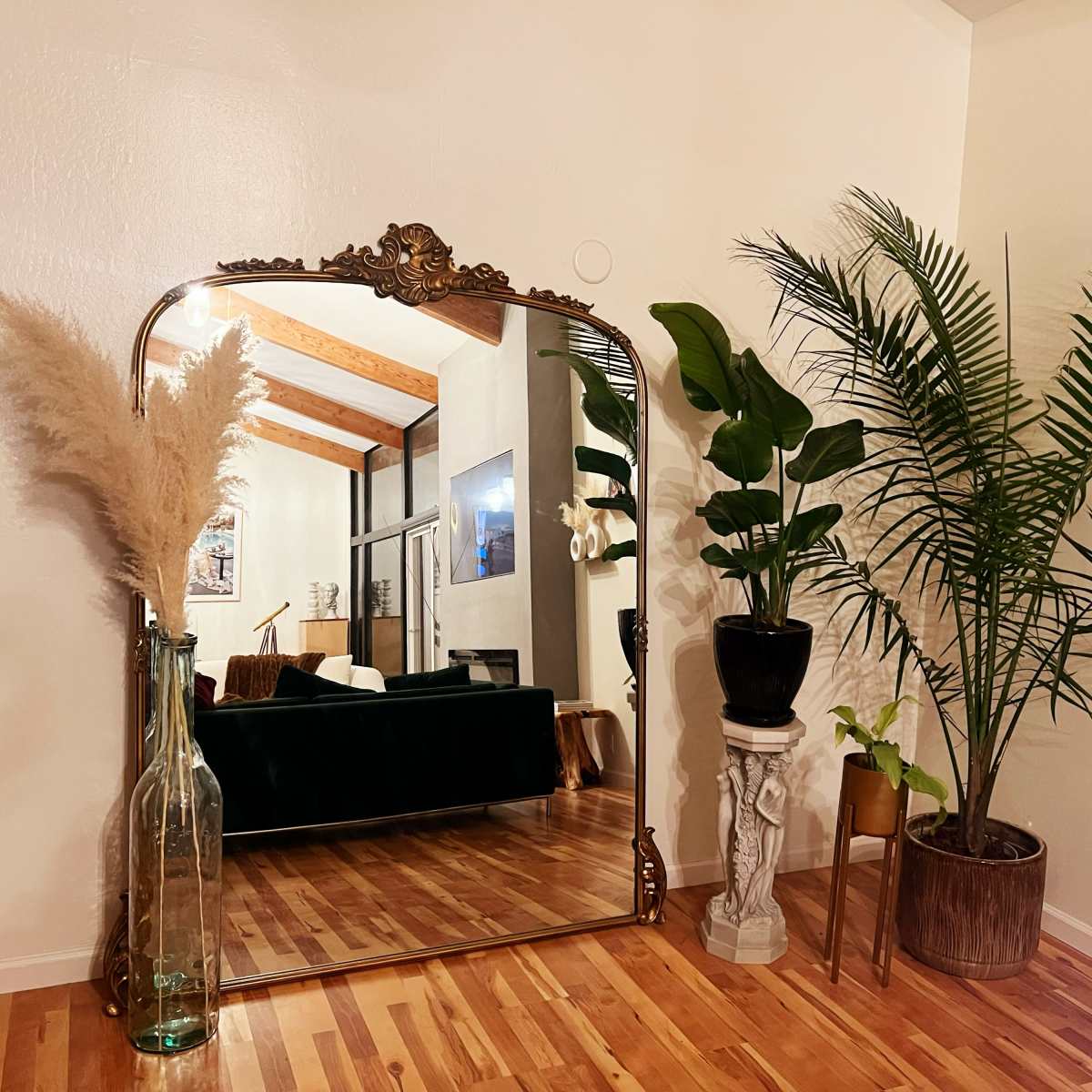 A large ornate mirror next to tall indoor plants and a decorative vase, with a wooden floor and a view of a living space in the background.