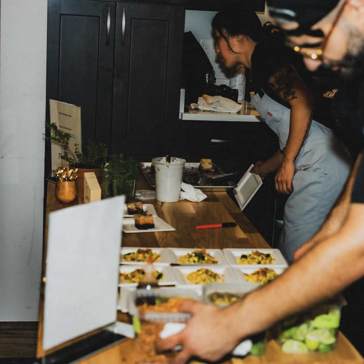 A person prepares food on a wooden table while another individual works in the background of a kitchen setting.