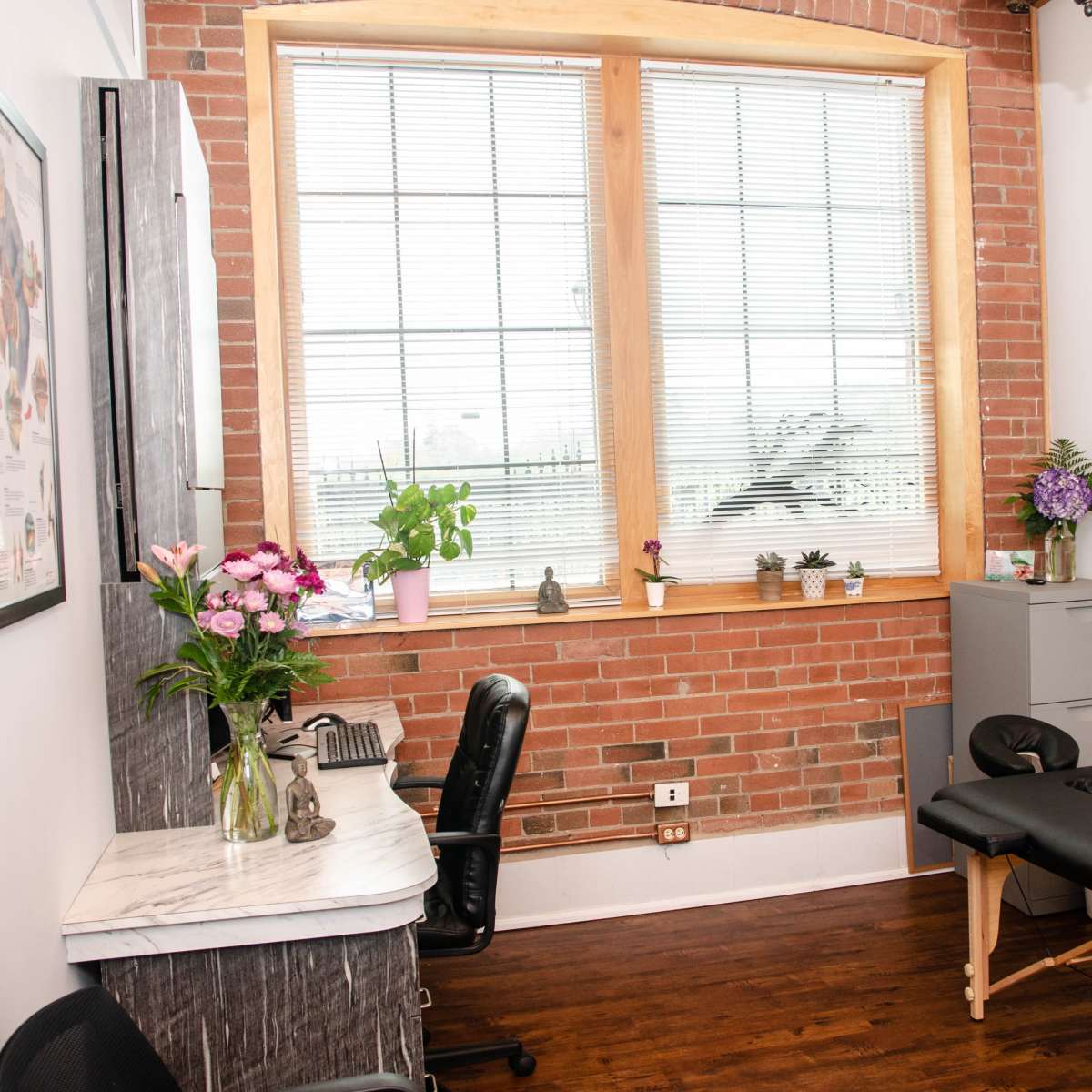A treatment room featuring a black massage table, a small desk with flowers, and a large window with blinds.
