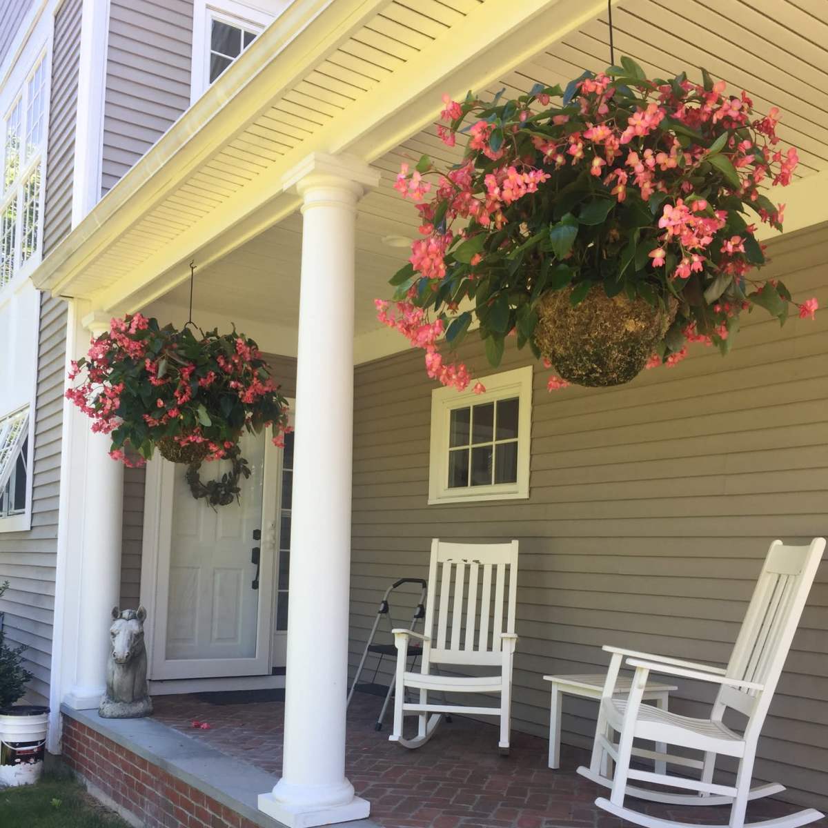 A front porch with two white rocking chairs, hanging floral baskets, and a decorative statue beside the entrance.